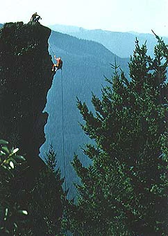 A rock climber hangs from a rope on the side of a tall, dark stone pinnacle, while another person sits on top of the formation; green trees and distant mountain ridges are visible in the background.