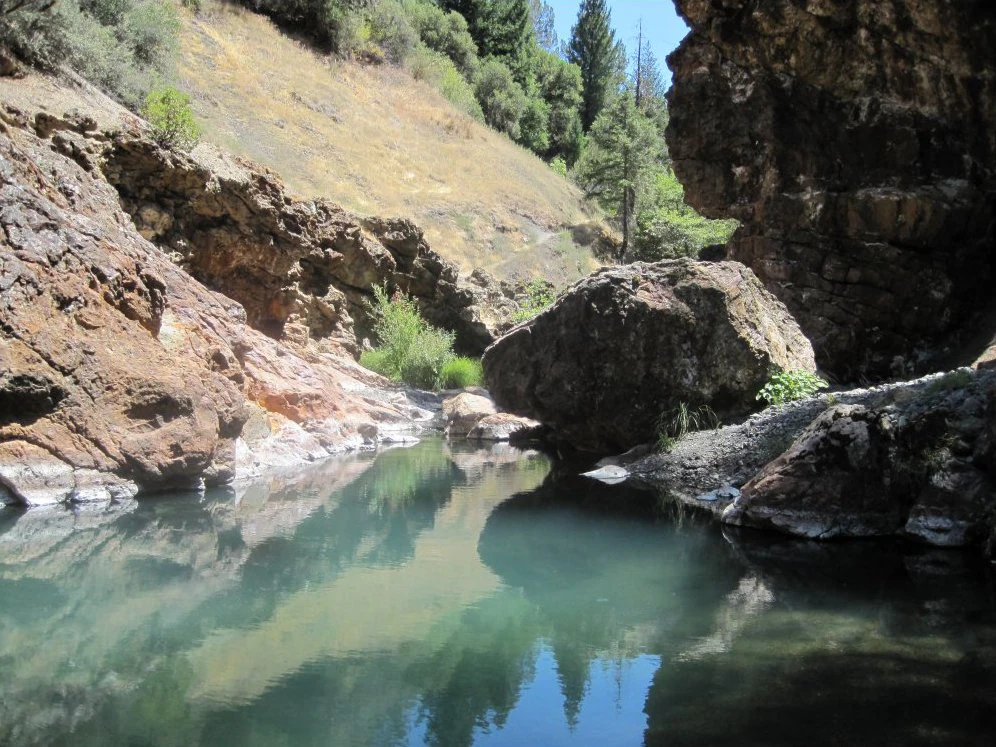A small pool of teal-colored water nestled between steep, rocky canyon walls and sparse coniferous trees.