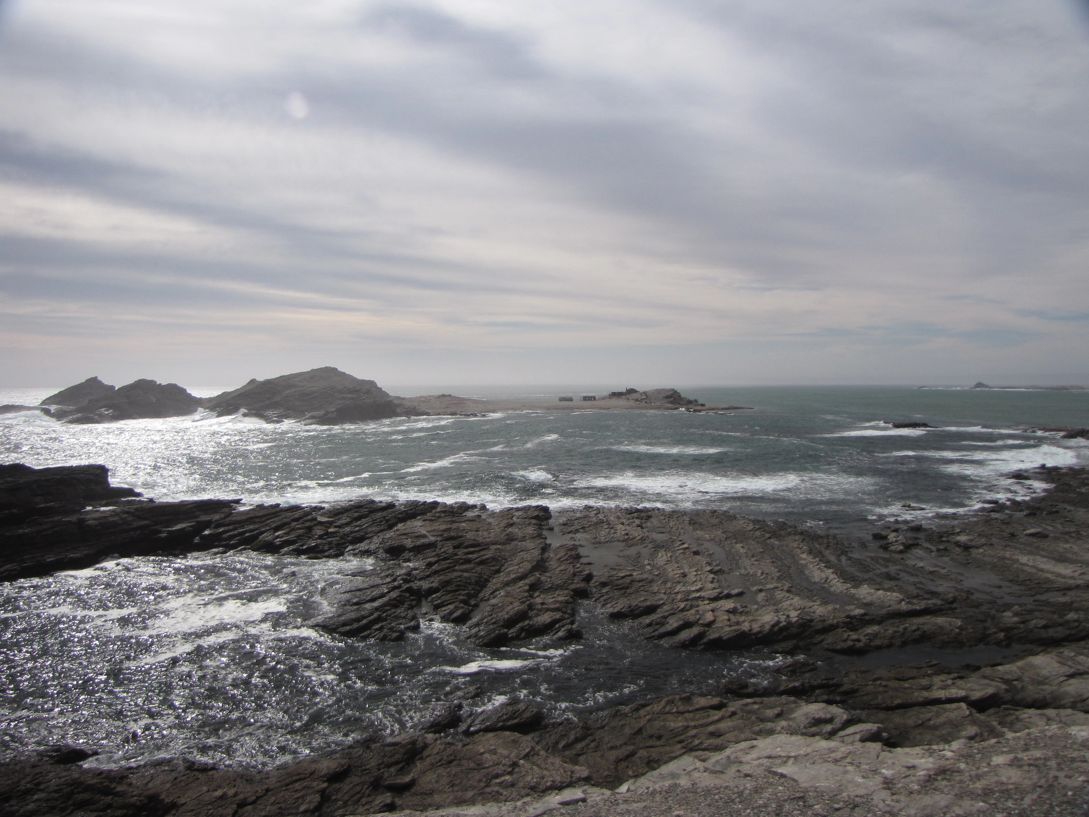 A wide view of a rocky coastline meeting the Atlantic Ocean under a cloudy, overcast sky, with the rocky silhouette of Halifax Island visible in the distance.