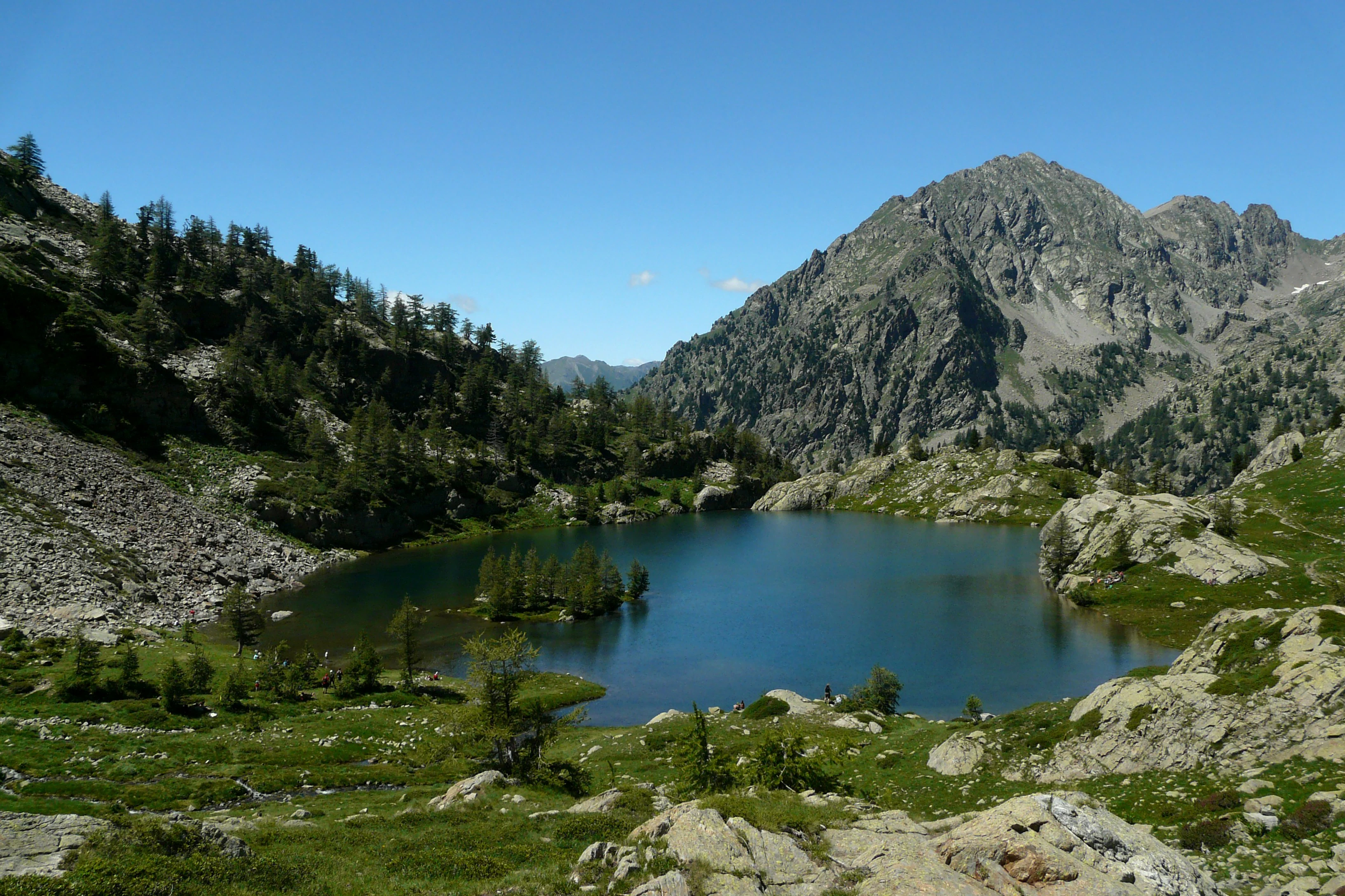A mountain lake surrounded by rocky terrain, green meadows, and forested slopes under a clear blue sky with a prominent mountain peak in the background.