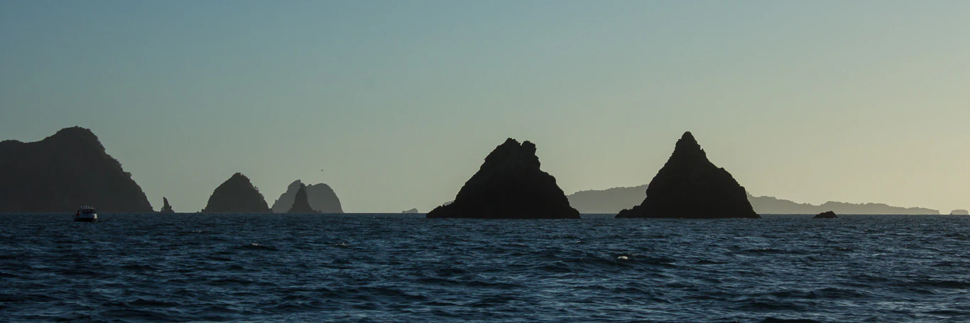 The silhouettes of several rugged islands and rock stacks are set against a bright, hazy sky above a dark blue ocean.
