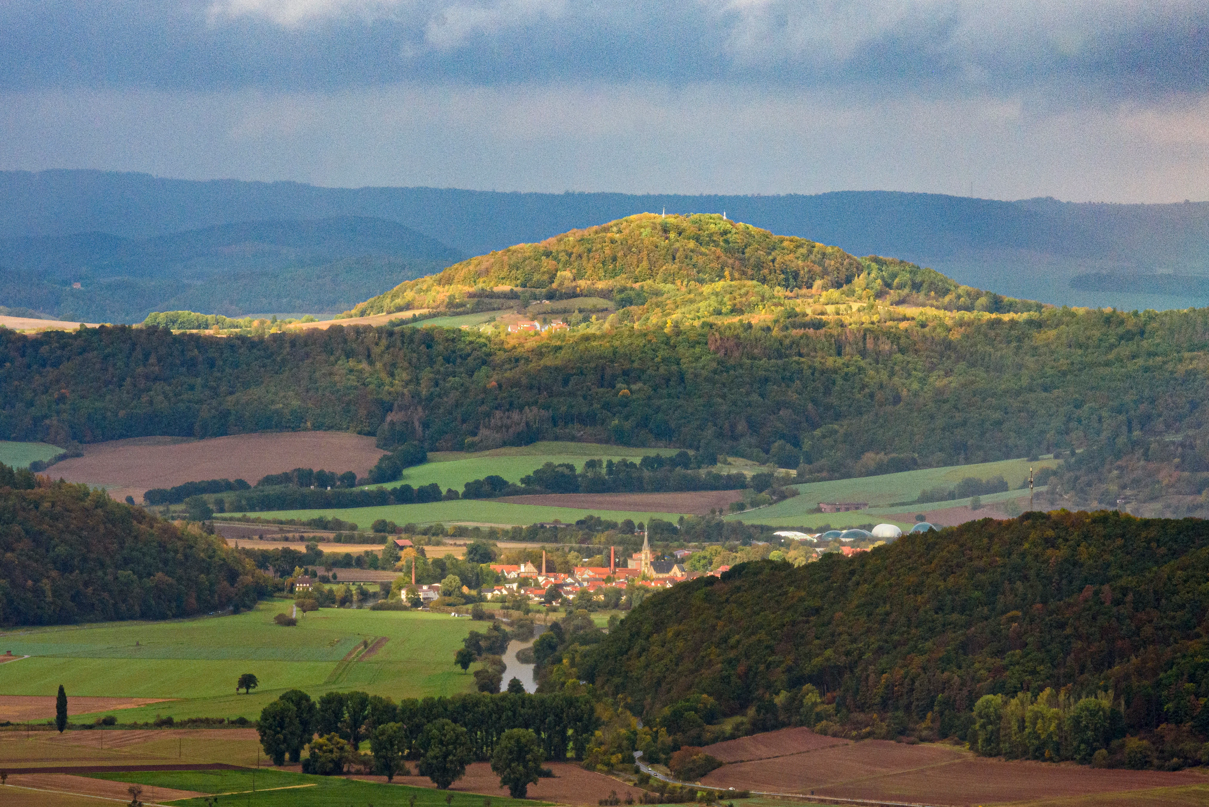 Una vista amplia del paisaje con colinas onduladas, laderas boscosas y tierras de valle agrícolas bajo un cielo nublado.