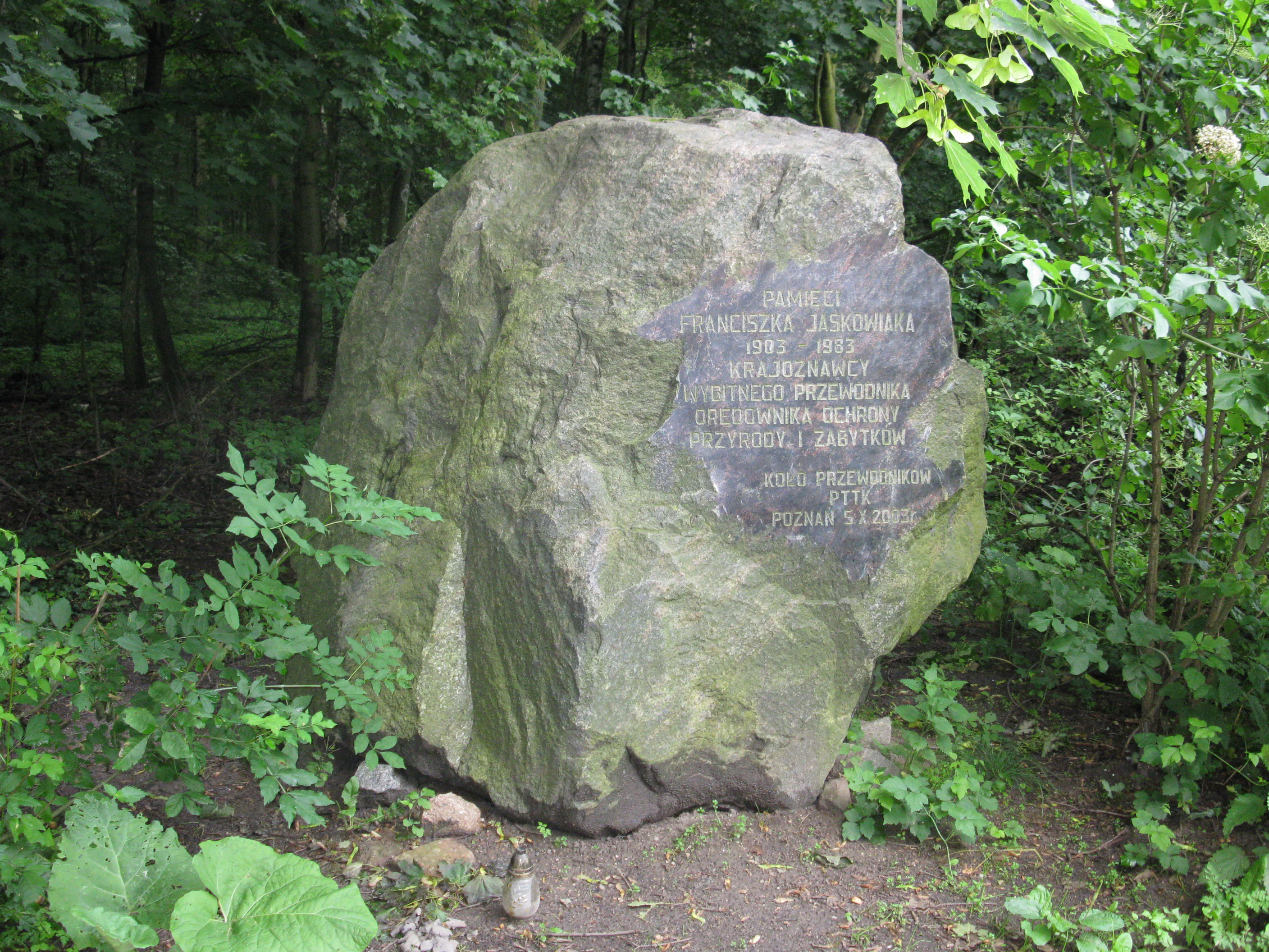 A large grey memorial boulder sits in a wooded area, featuring an inscribed metal plaque dedicated to Franciszek Jaskowiak.