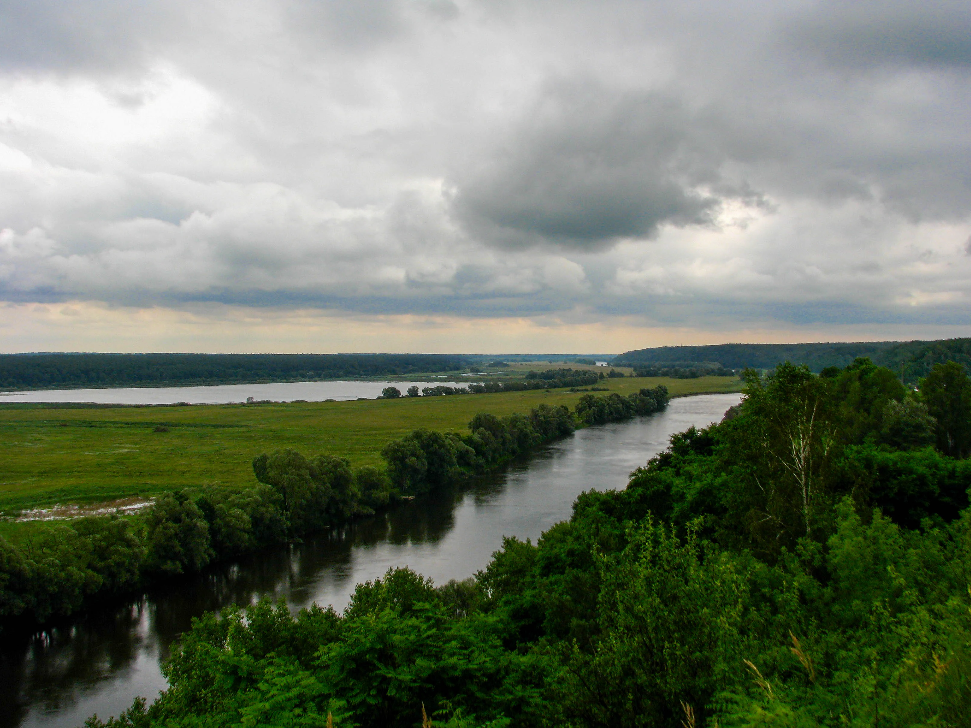 Wide landscape view of a river valley with green fields on one side and forested hills on the other under an overcast sky