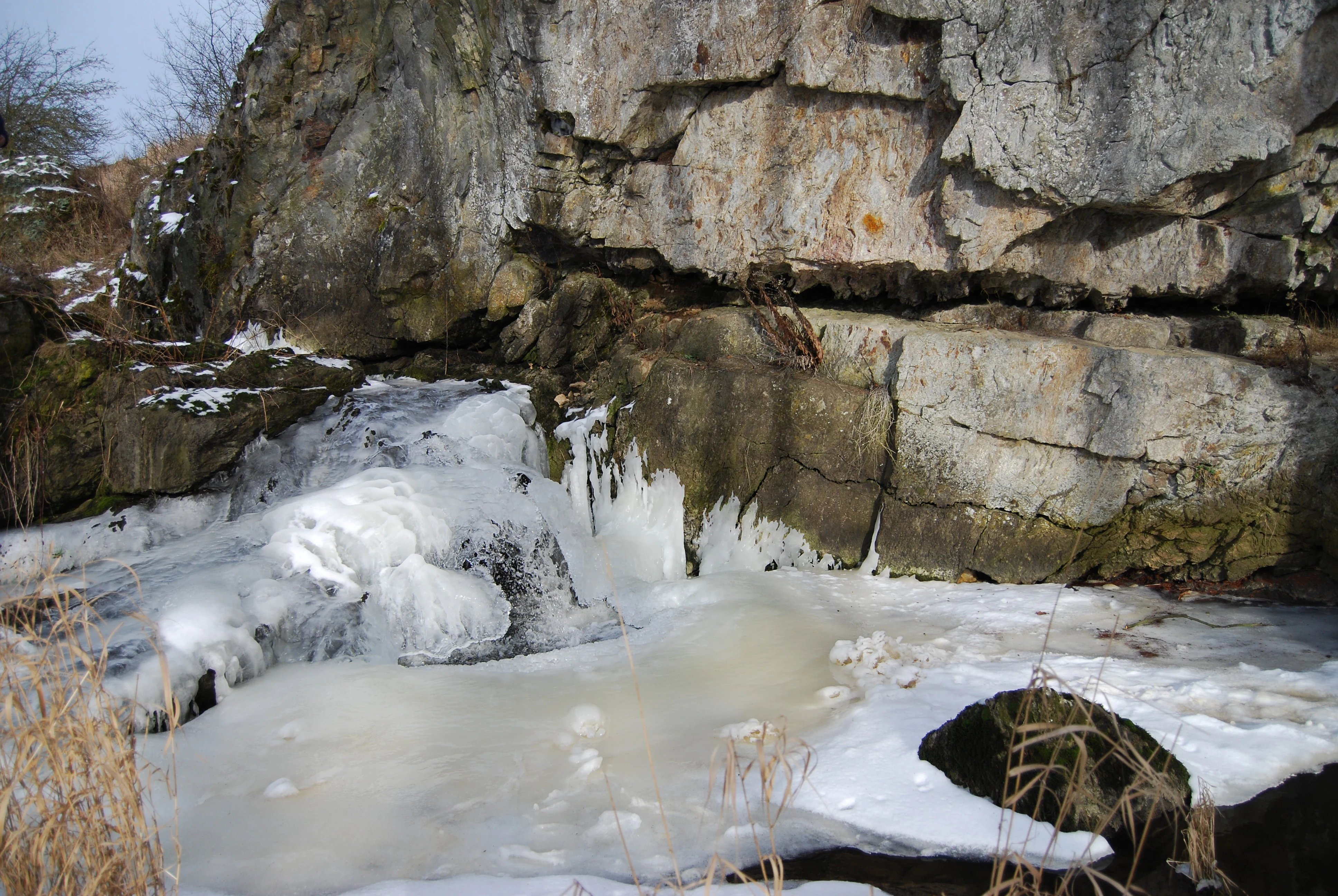 A partially frozen stream with icy rapids and icicles flowing past large rocky cliffs, with snow and dry grass in the foreground.