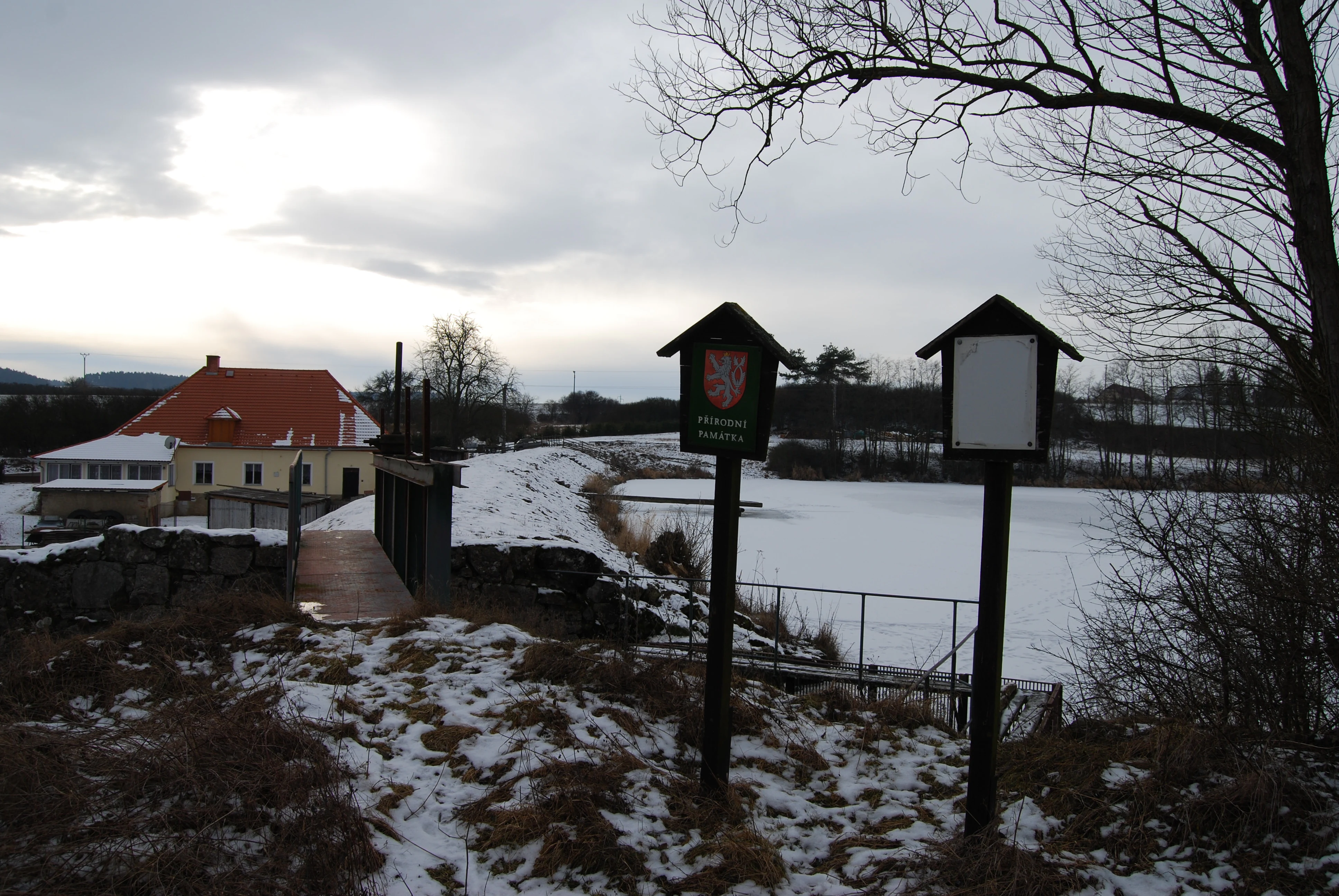 Two wooden signposts with roofed tops stand in the foreground on a snowy bank, with a frozen snow-covered pond, a yellow building with a red roof, bare trees, and an overcast sky in the background