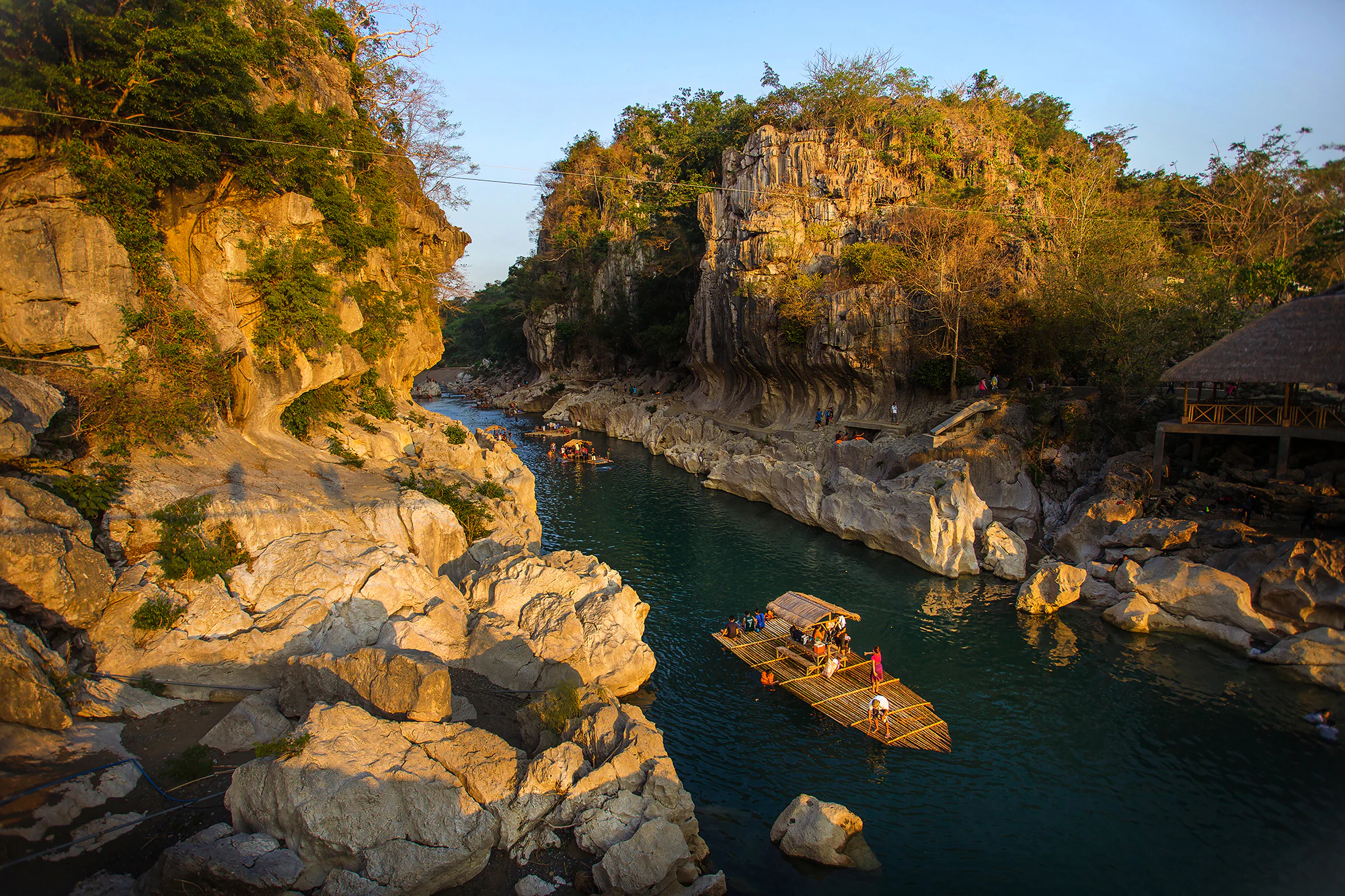 A river flowing between rocky limestone cliffs with a bamboo raft carrying people, surrounded by forested hills under a clear sky