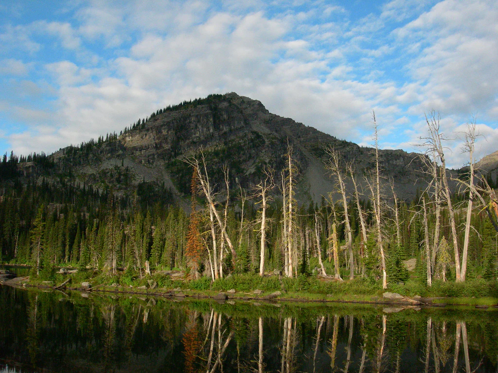 A calm mountain lake reflects the surrounding evergreen forest and the steep, rocky slopes of a mountain peak under a partly cloudy sky.
