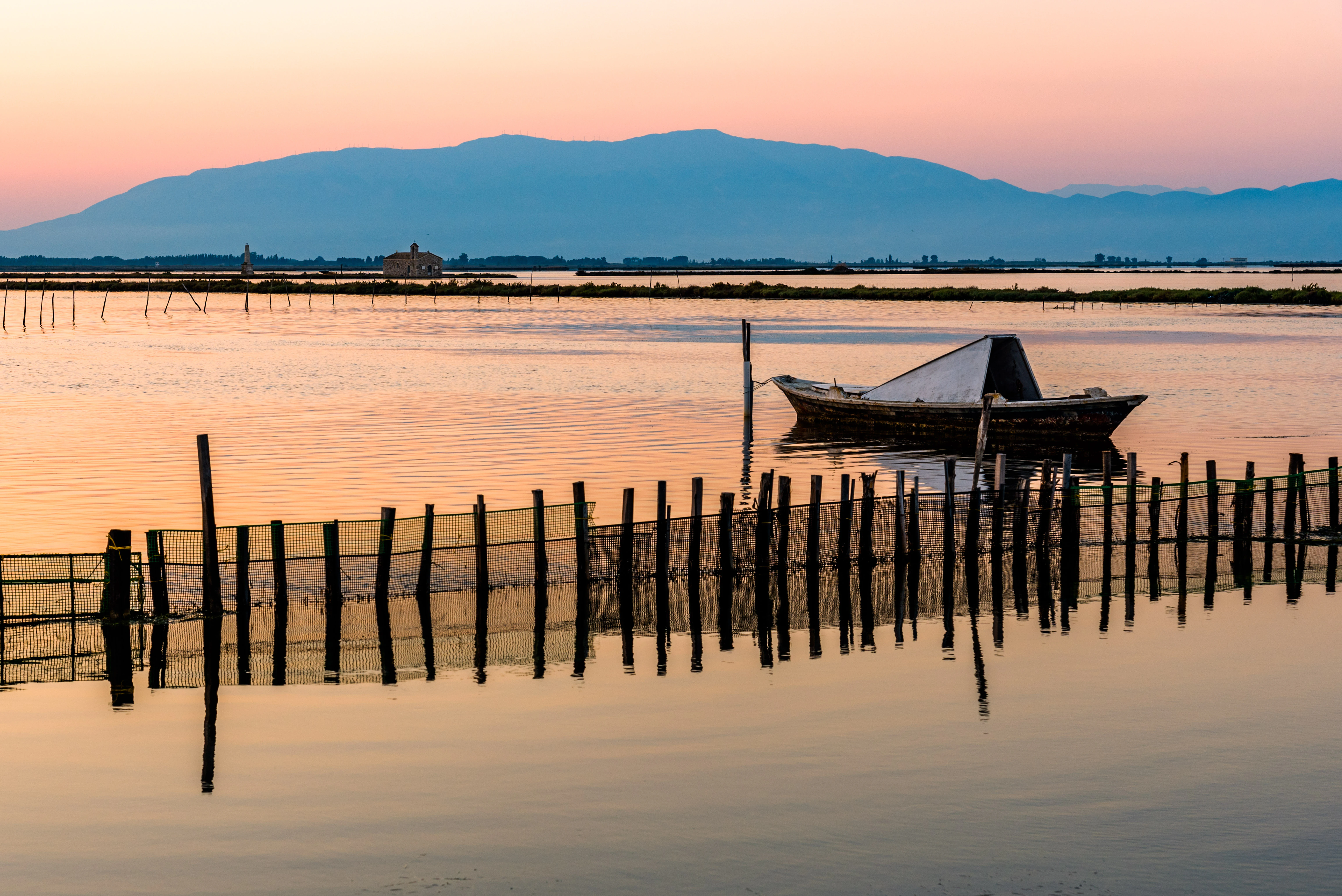 Un pequeño barco de pesca está amarrado en una laguna tranquila y plana al atardecer. Una hilera de postes de valla de madera se alza en primer plano, reflejándose en el agua, con montañas onduladas en la distancia brumosa.
