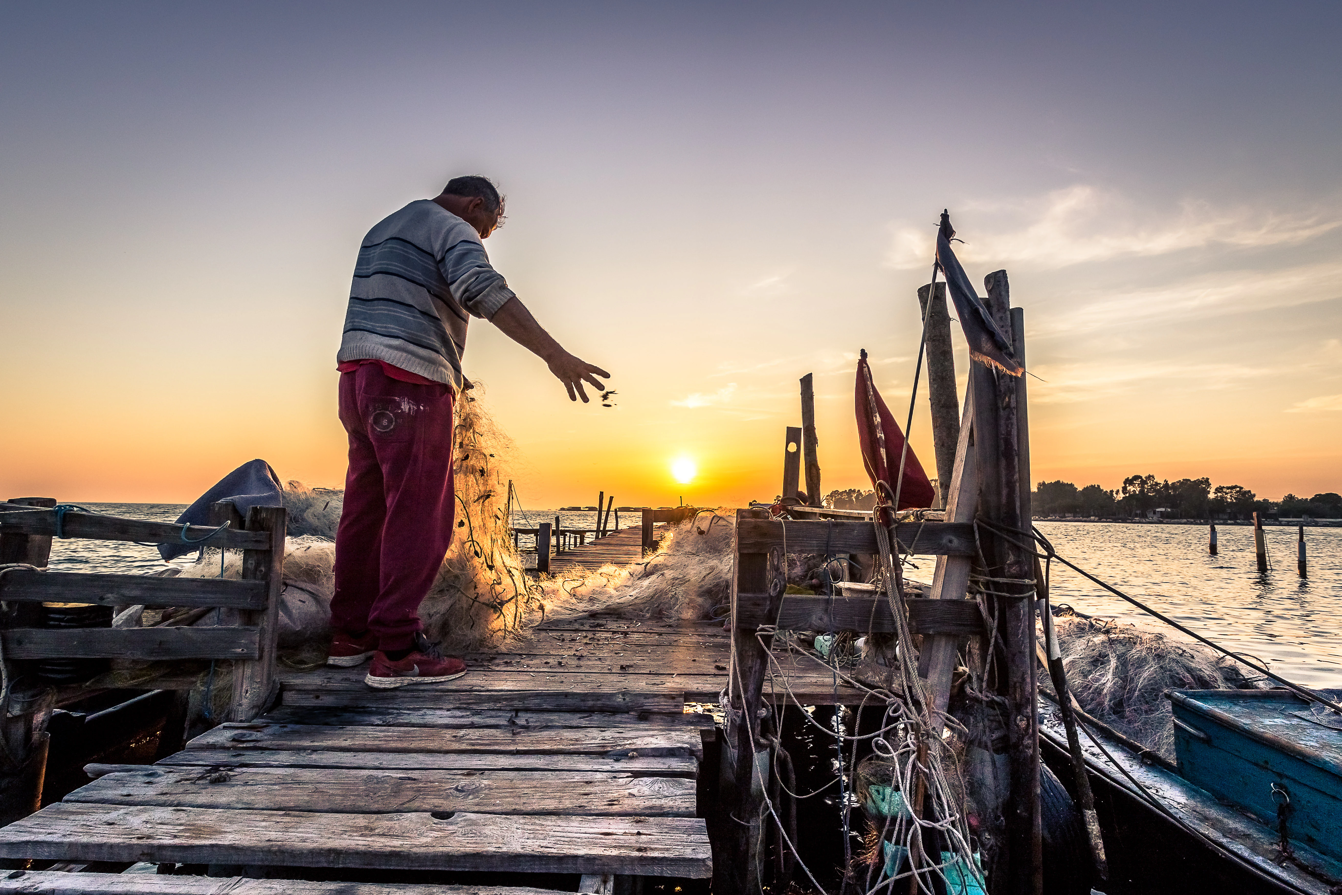 Un pescador silueteado con ropa informal lanza una red de pesca desde un muelle de madera desgastado al agua al atardecer.