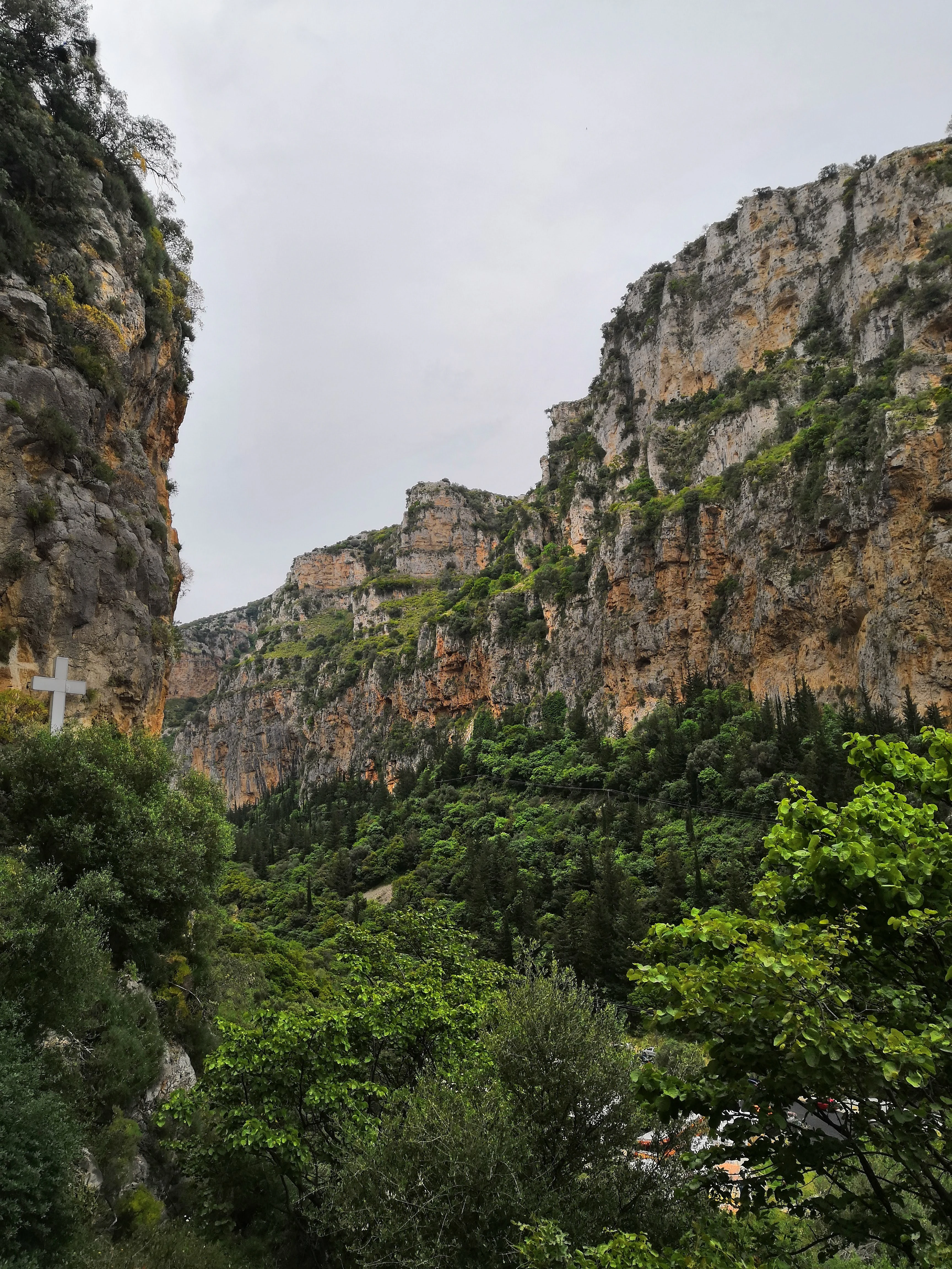 Un desfiladero escarpado de piedra caliza con acantilados altos y dentados cubiertos de parches de vegetación, con una prominente cruz blanca visible en el lado izquierdo, cerca de la pared rocosa.