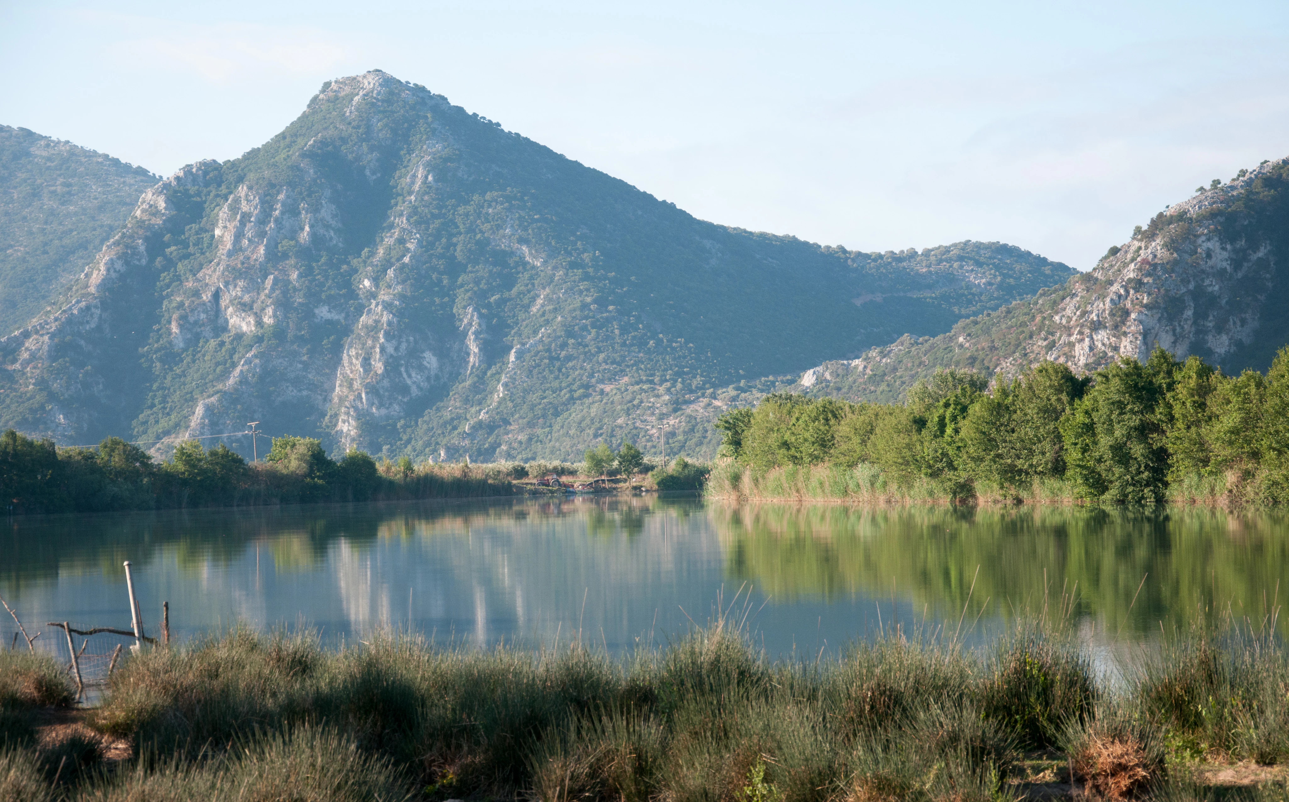Una vista amplia de un río tranquilo que refleja montañas rocosas al fondo, con vegetación ribereña verde y frondosa a lo largo de las orillas.