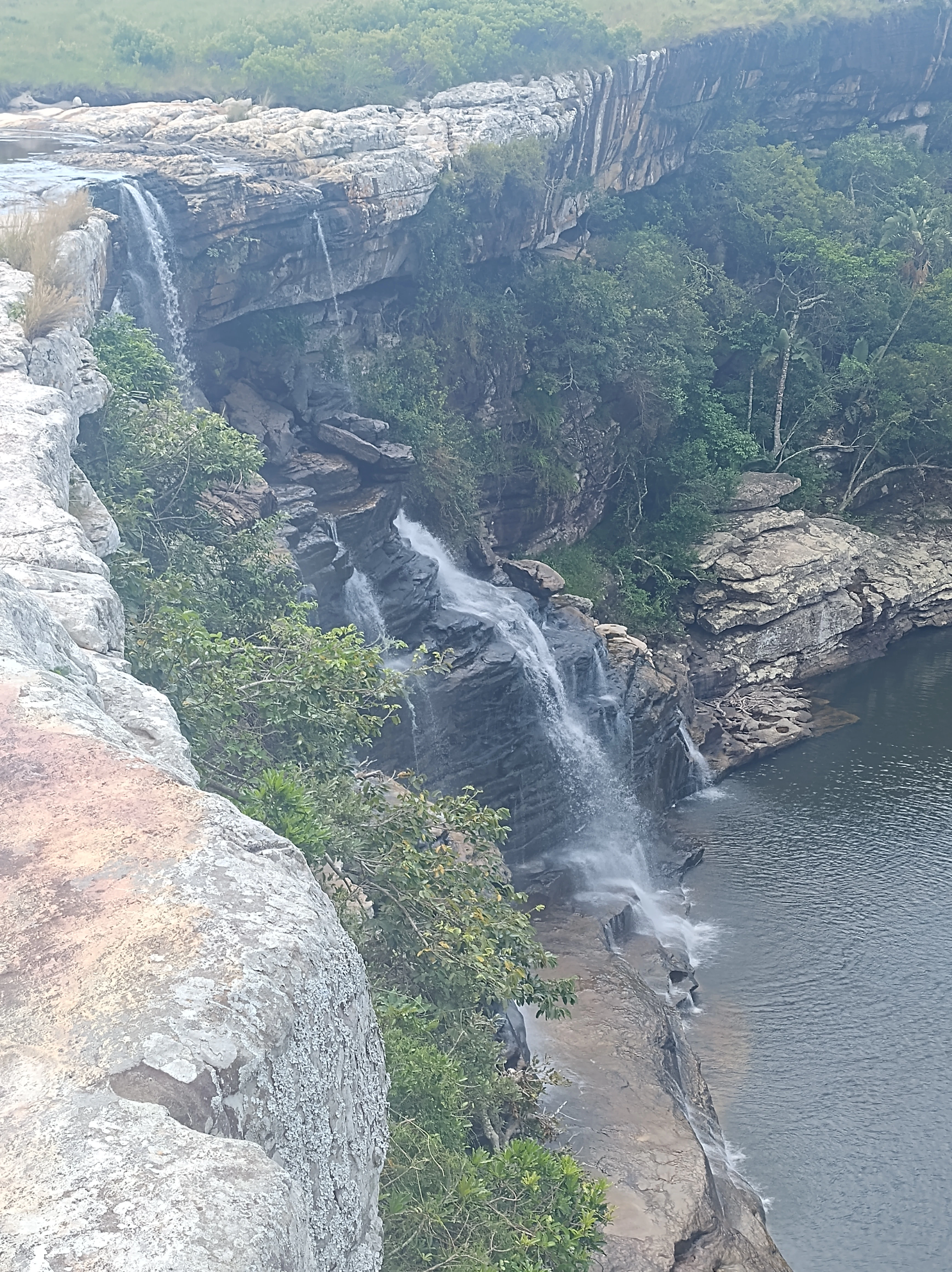 A waterfall flows over a layered rocky cliff into a pool below, surrounded by green coastal grassland and forest vegetation.