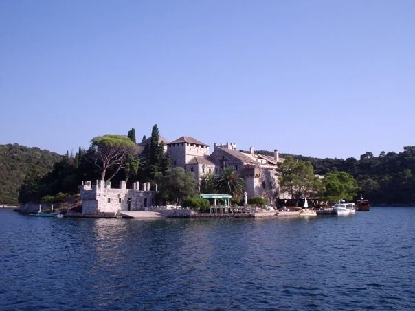 A historic stone building complex on a small island surrounded by calm blue water with trees and hills in the background