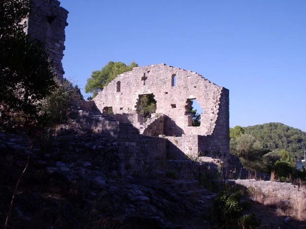 Stone ruins of an early Christian basilica with open walls and windows, set against a clear blue sky with trees and rocky terrain