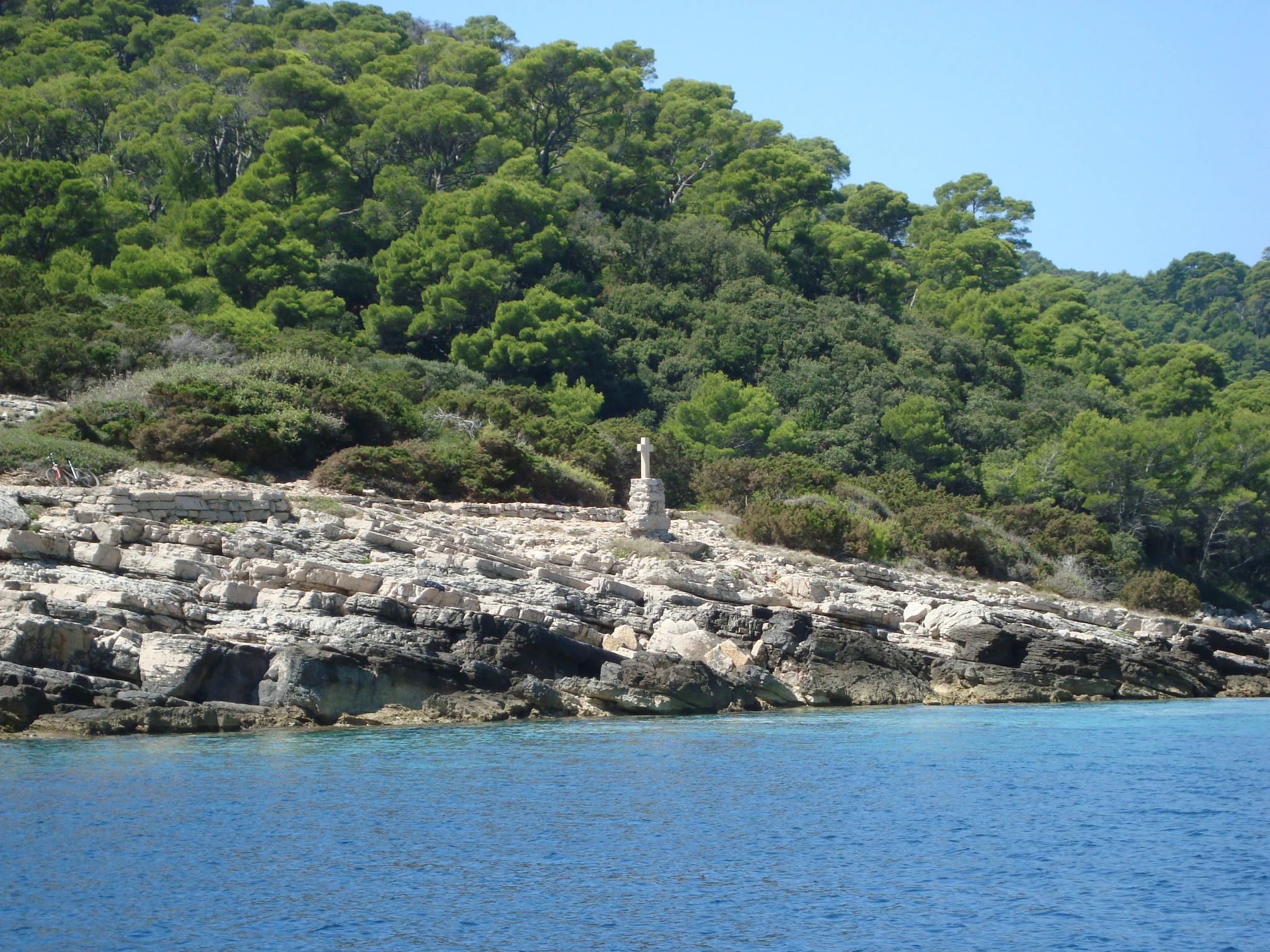 Rocky shoreline with stone cross overlooking blue water and forested hills