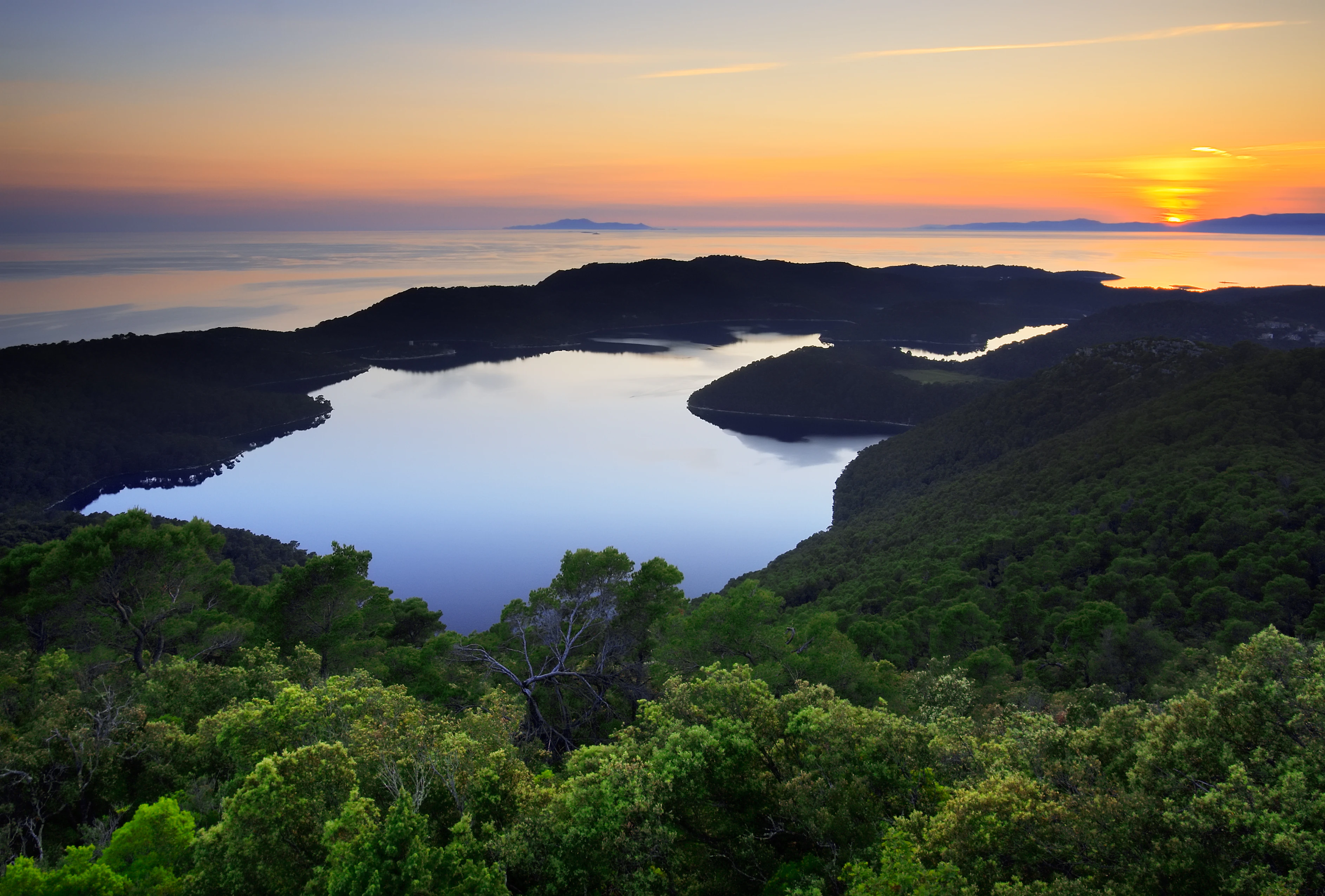 Sunset over a lake surrounded by dense forest with distant hills under an orange and blue sky