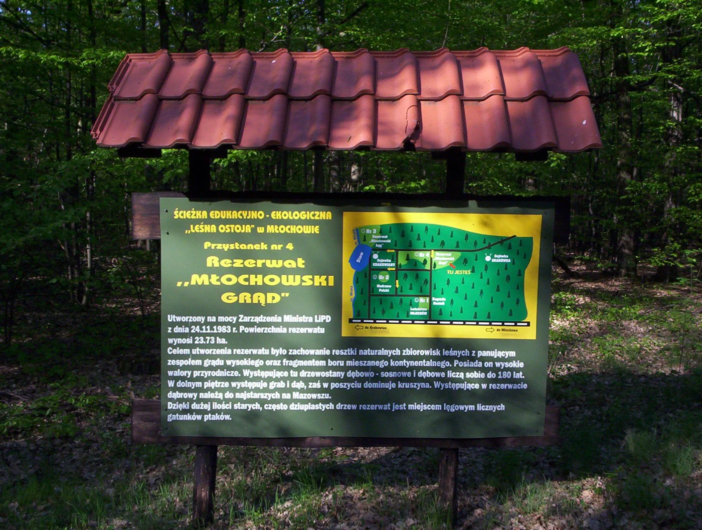 A rectangular outdoor educational information sign with a red tile roof, standing in a forest. The sign features Polish text and a small green map of the nature reserve.