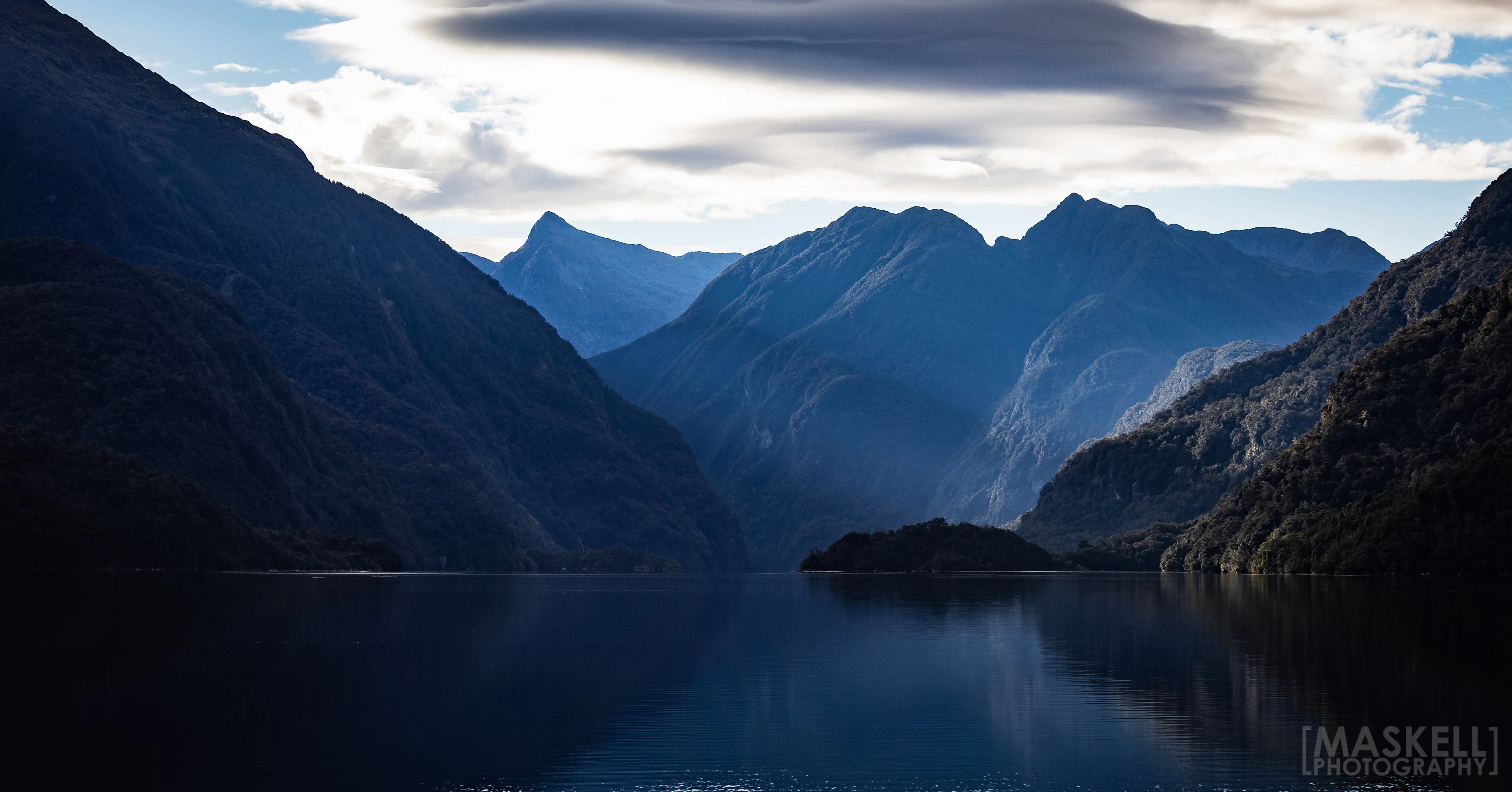 A wide, landscape view of dark, steep mountain slopes rising from a calm, reflective body of water under a dramatic, clouded sky.
