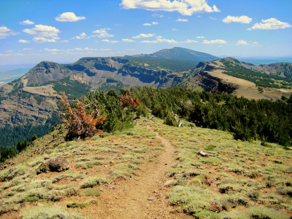 A dirt hiking trail winding across a grassy mountain ridge with a vast volcanic mountain range in the background under a blue sky.