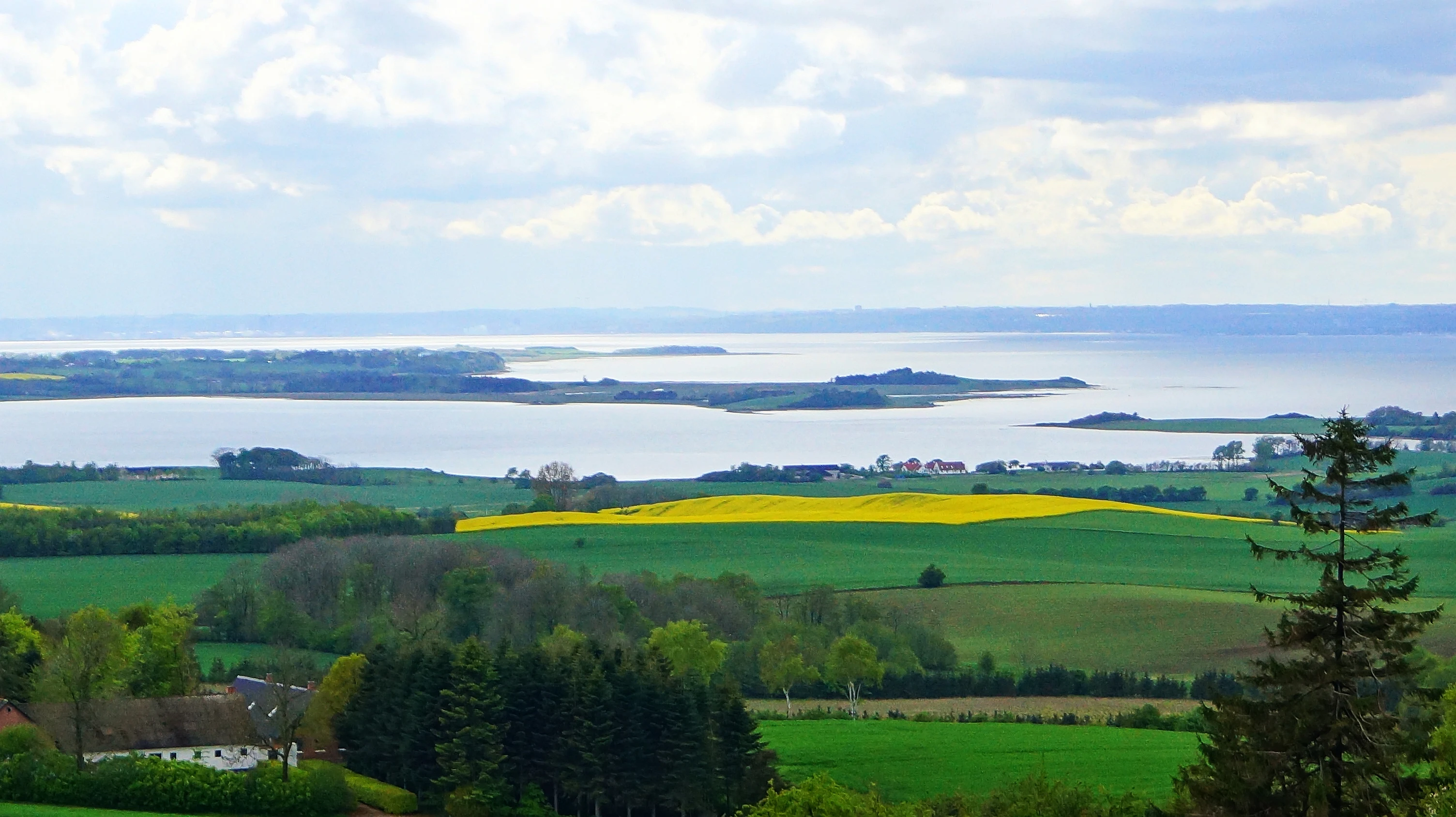 A wide, elevated landscape view featuring rolling green fields, patches of yellow crops, forests, and a large body of water in the background under a cloudy sky.