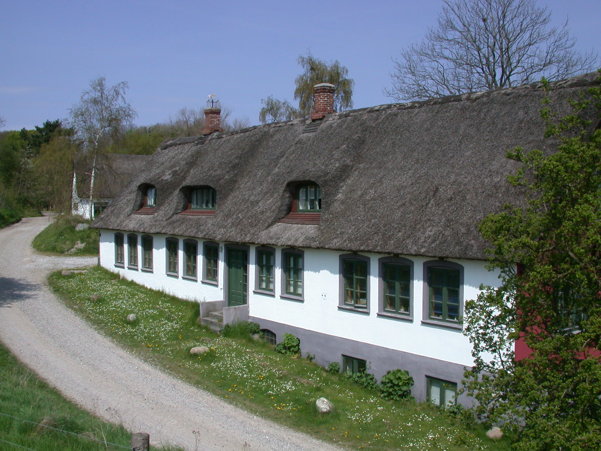 Traditional white house with thatched roof on grassy slope beside gravel road, trees in background