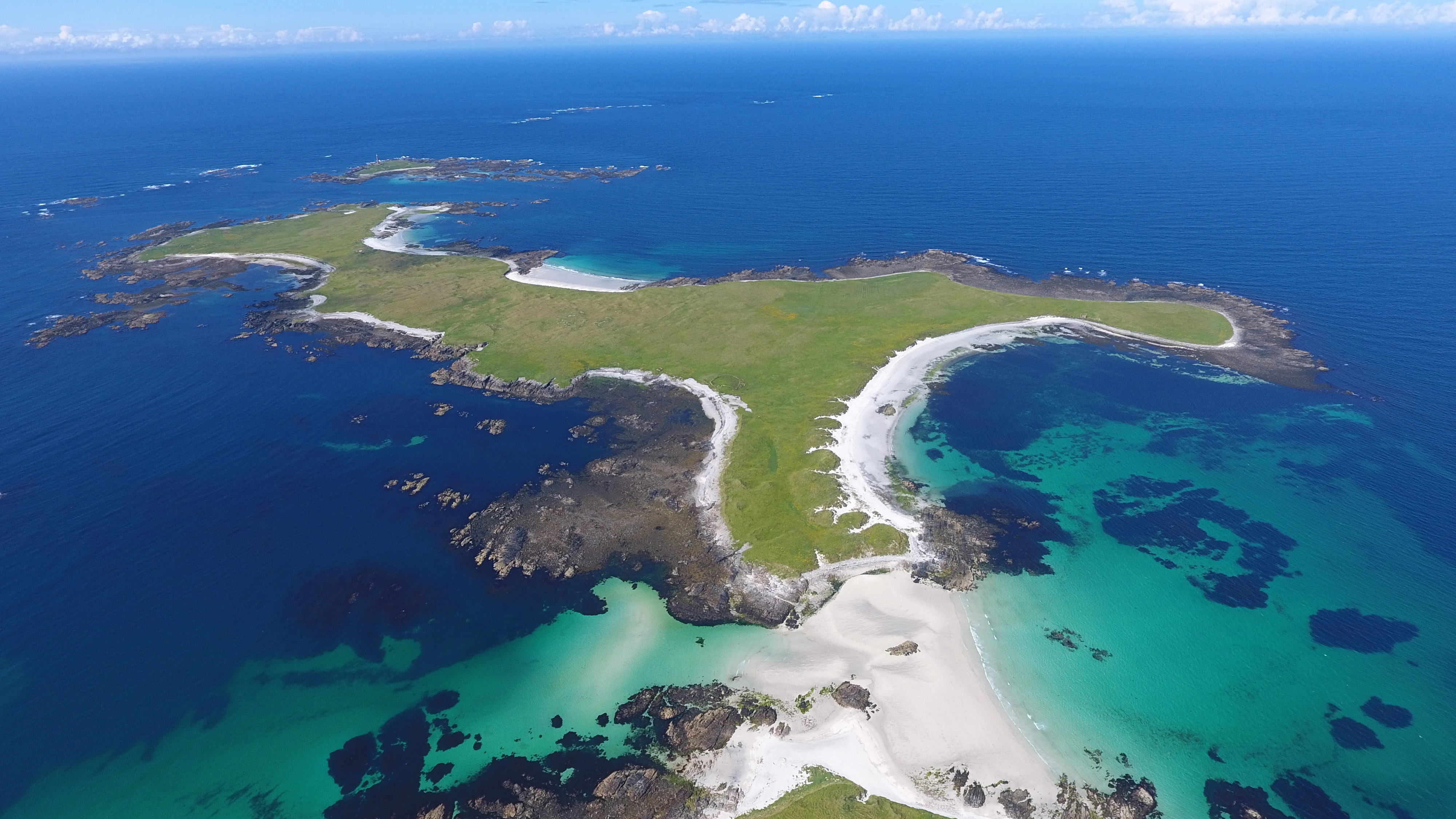 An aerial drone view of the remote, green, and flat Monach Islands surrounded by clear turquoise and deep blue ocean waters.