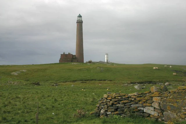 A tall, slender stone lighthouse stands on a grassy hill, with a small, modern white lighthouse tower nearby and a dry stone wall in the foreground.