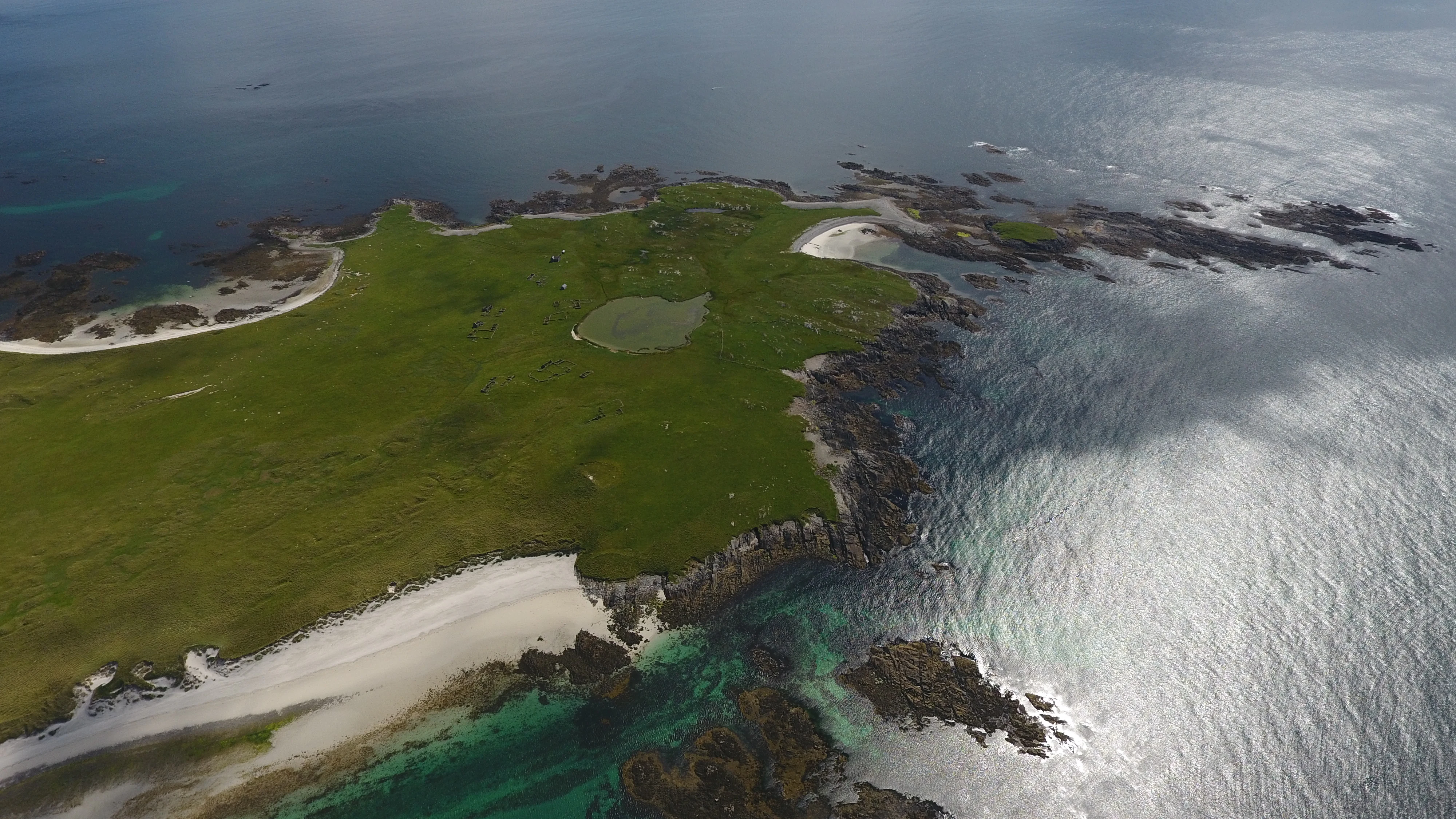 A high angle aerial drone view of the Monach Islands featuring a bright green, grassy island surrounded by blue sea, with visible white sand beaches, rocky shorelines, and a small circular inland pool.