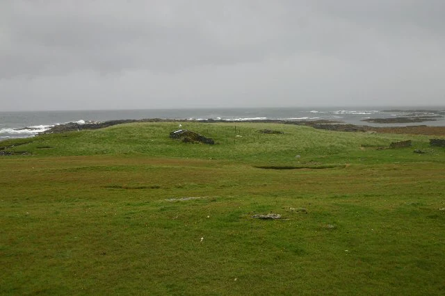 A wide, low-angle shot of a grassy, treeless coastal landscape under a gray, overcast sky, featuring a small, crumbling stone ruin in the midground and the ocean in the background.