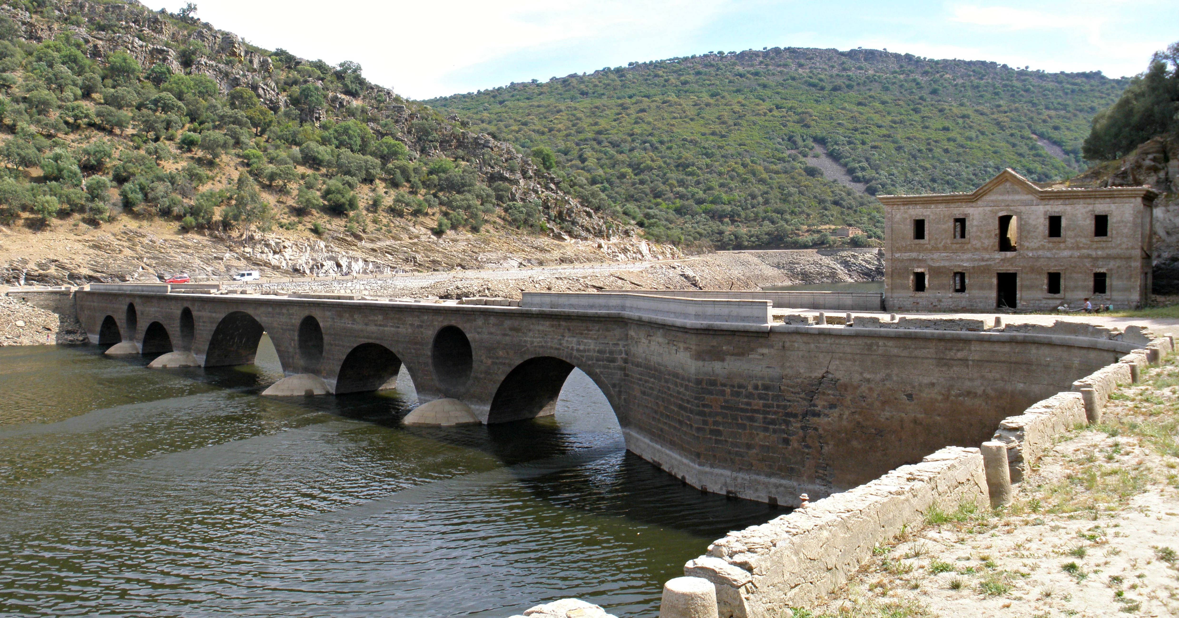 Stone bridge with multiple arches spanning a body of water, with hills and a building in the background