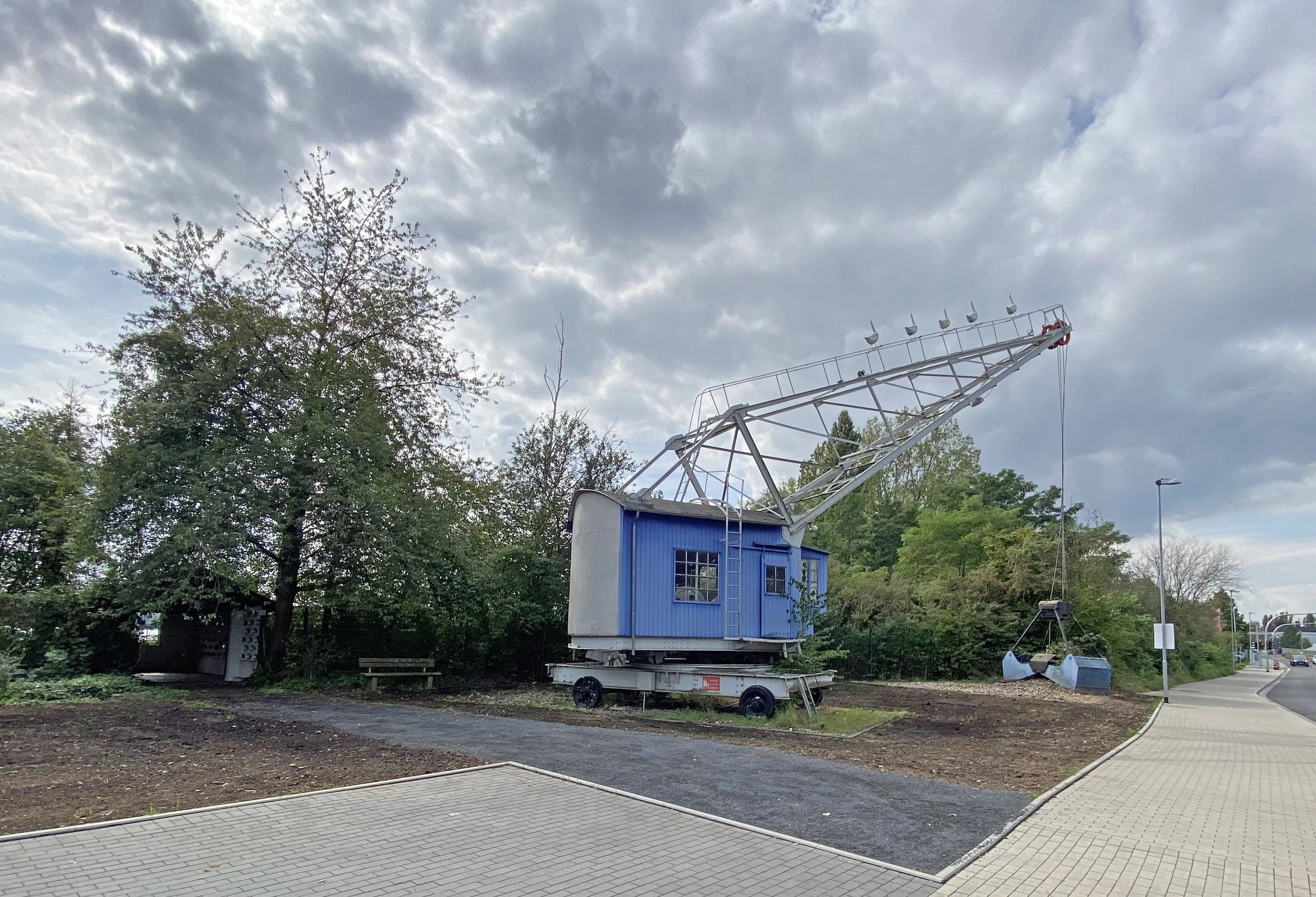 A vintage blue and white industrial gravel crane standing on wheels on the edge of a paved path in a green area under a cloudy sky.