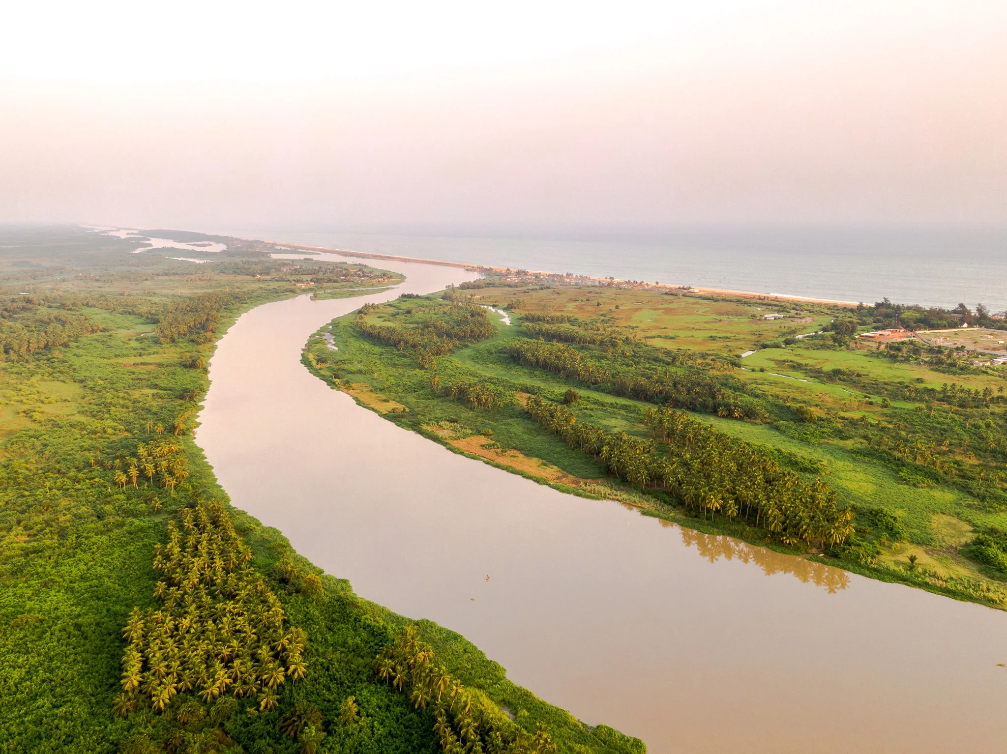 Aerial perspective looking over the wide, calm Mono River, flanked by dense green vegetation and palm trees, leading toward the distant horizon where the river meets the coastline.