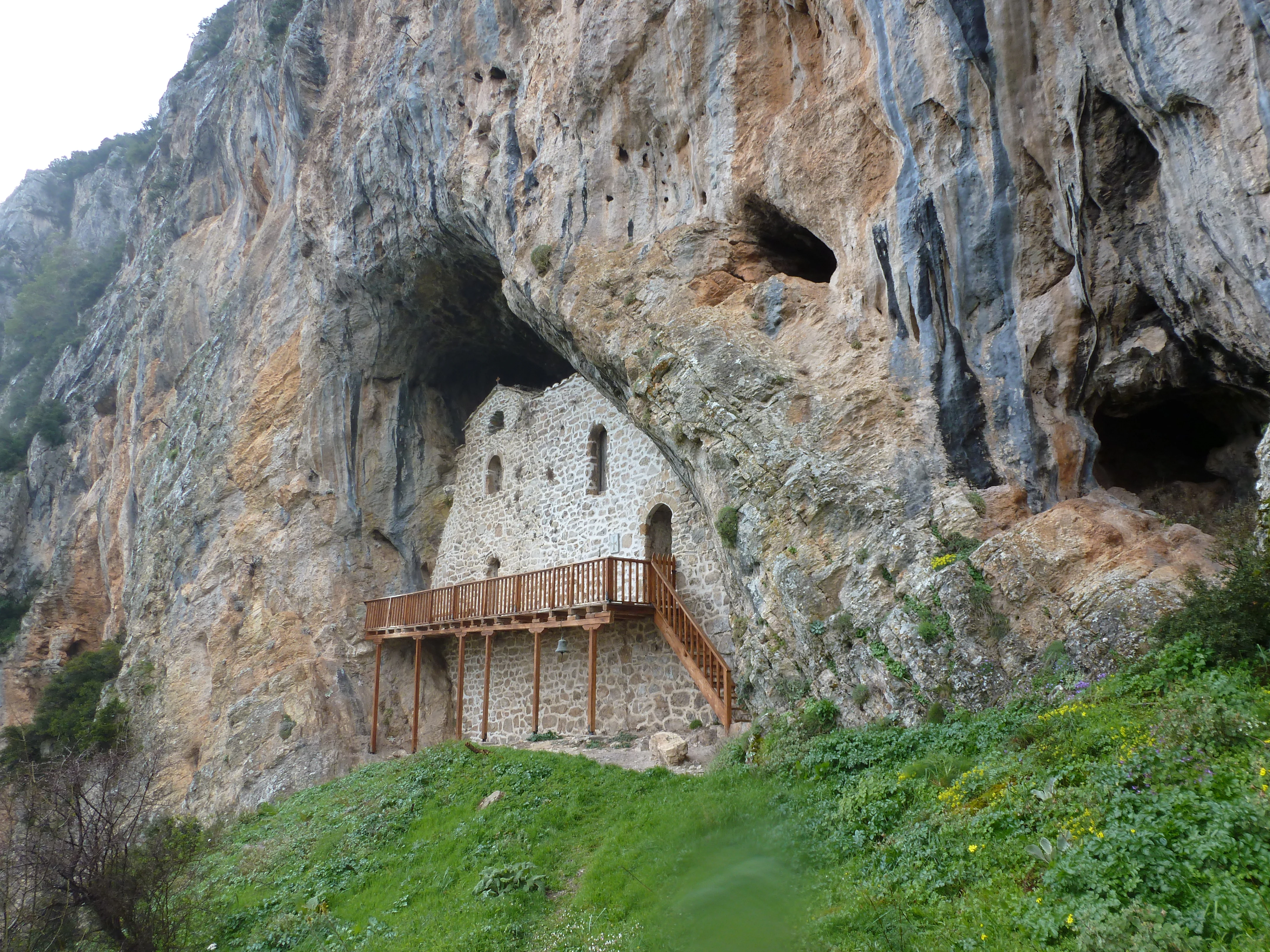 A small stone building constructed into a large natural cave opening in a cliff face, with a wooden balcony and stairs, surrounded by grassy slopes.