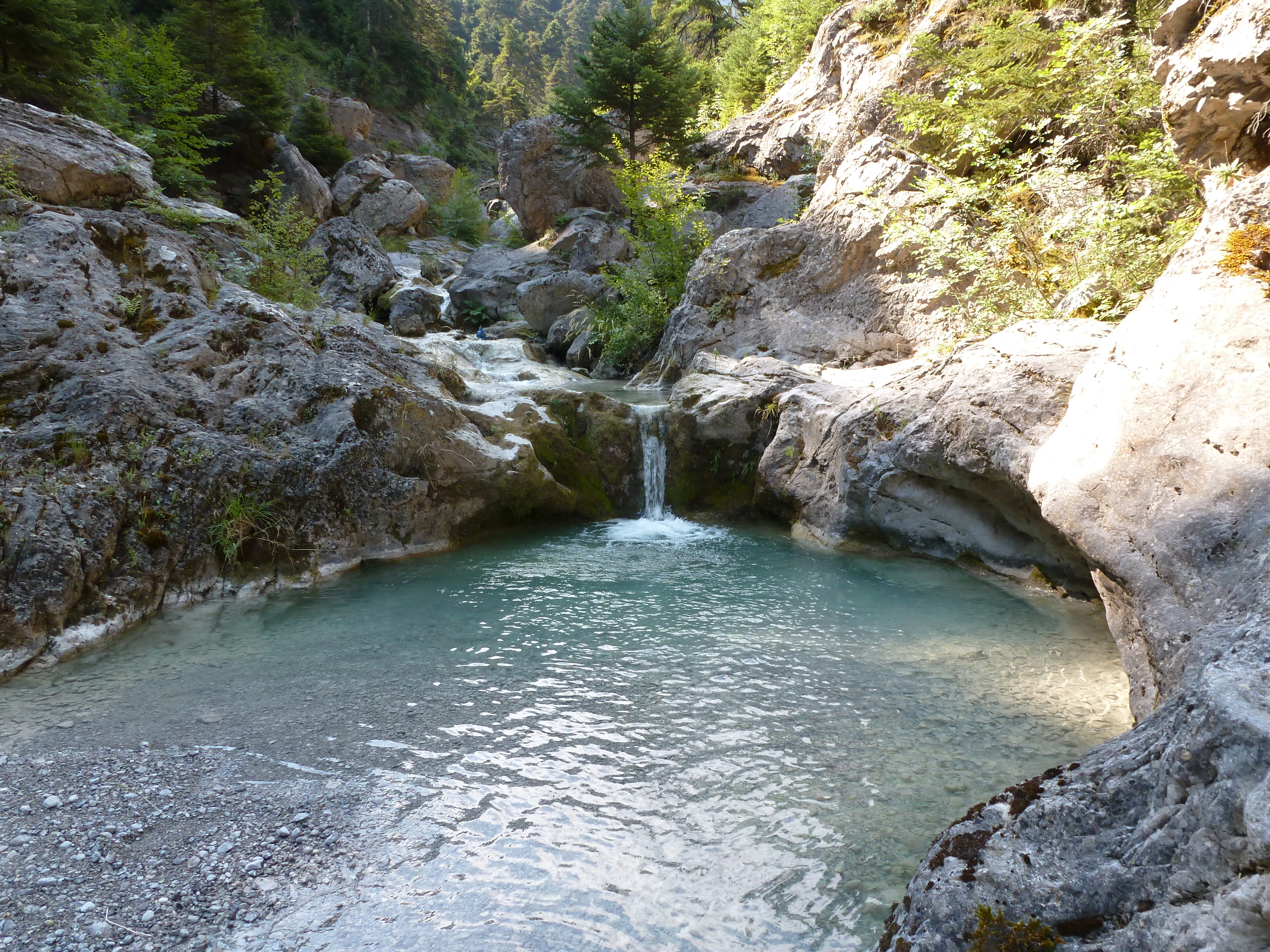 A small mountain stream flows over rocky terrain into a turquoise-colored pool, surrounded by rugged cliffs and evergreen trees.