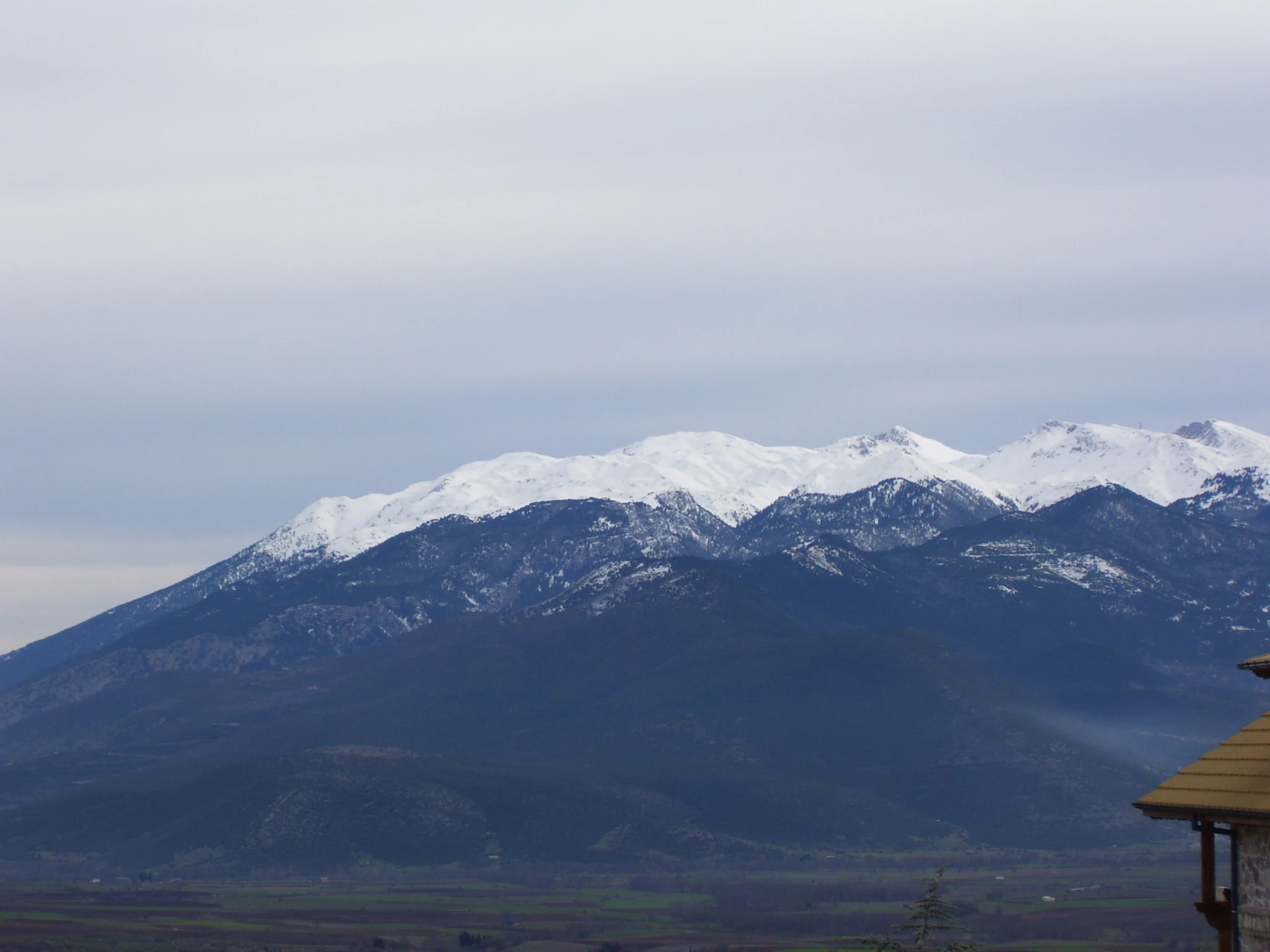 A wide view of the dark, forest-covered slopes of Mount Parnassus topped with snow against a cloudy sky, with the roof of a building partially visible in the lower right.