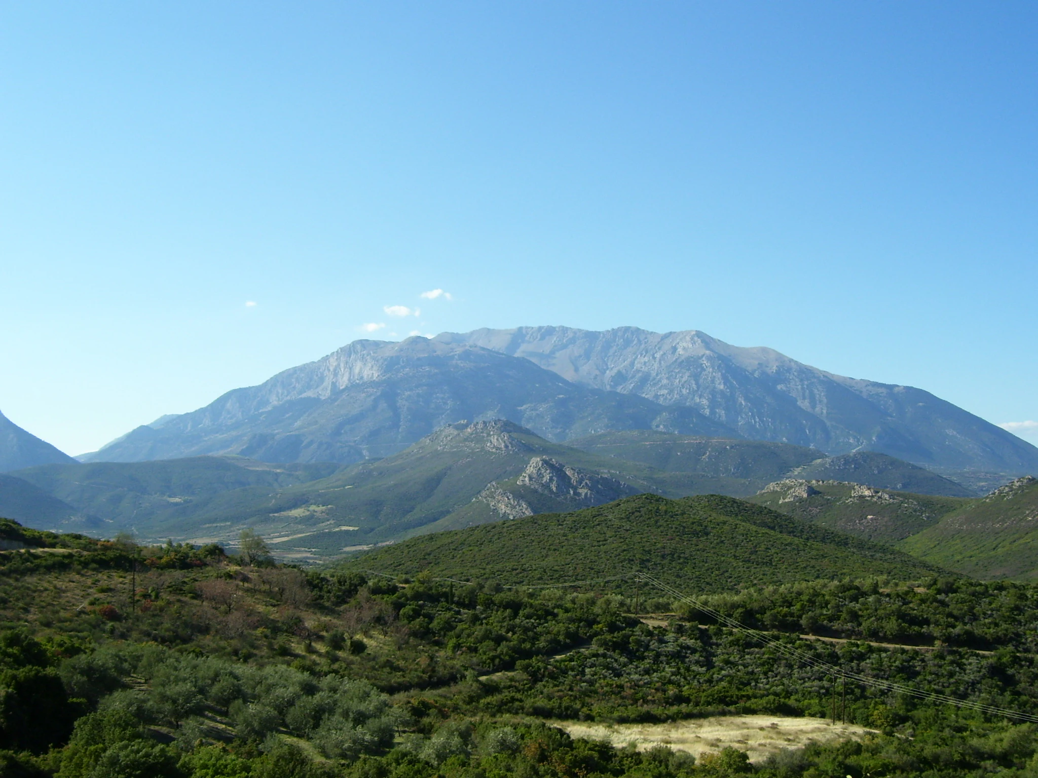 A wide, eye-level landscape view of the rocky, grey summits of Mount Parnassus rising above rolling, green forested slopes under a clear blue sky.