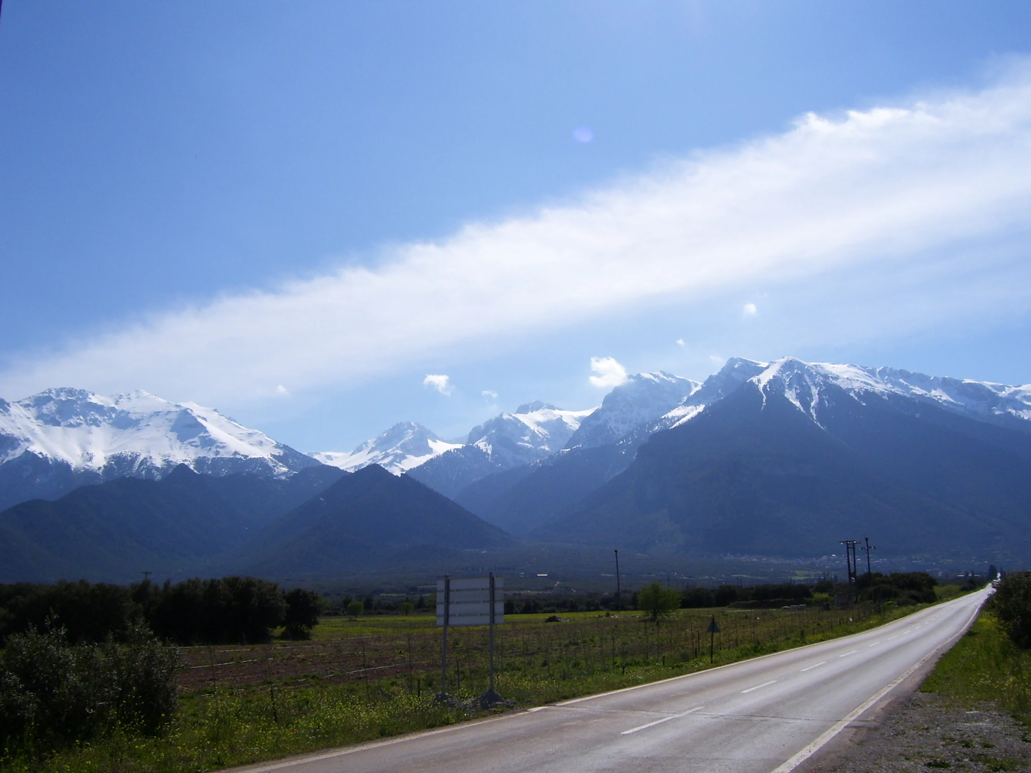 A wide, eye-level shot of the snow-capped Parnassos mountains rising behind a flat, rural landscape with a paved road leading toward them under a clear blue sky.