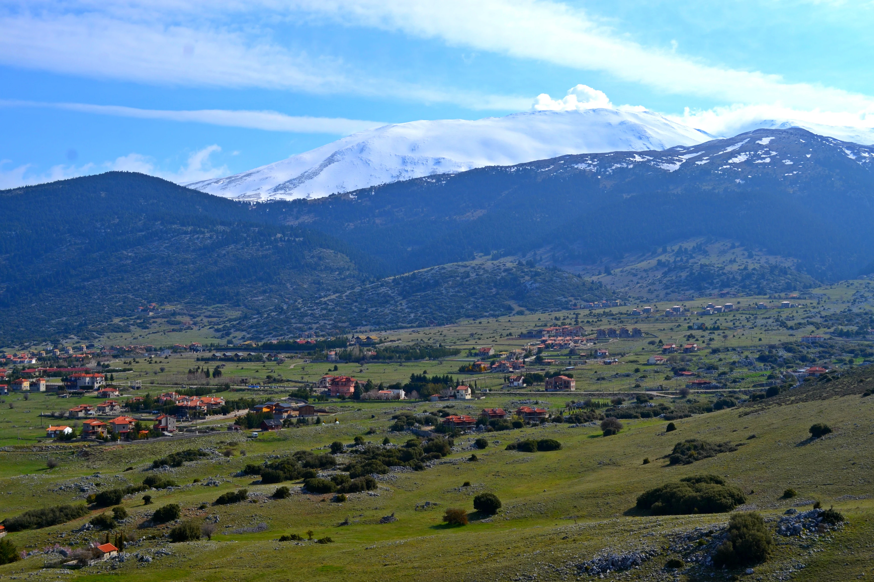 A wide view of a grassy plateau with small stone buildings of the Kalivia settlement, backed by the snow-capped peak of Mount Parnassus under a blue sky.