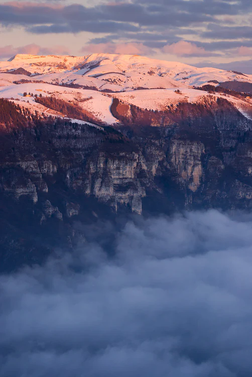 A wide, vertical shot of a mountain range covered in snow at sunset, with the summit glowing in pink light and the valleys below filled with a thick layer of blue-toned clouds.