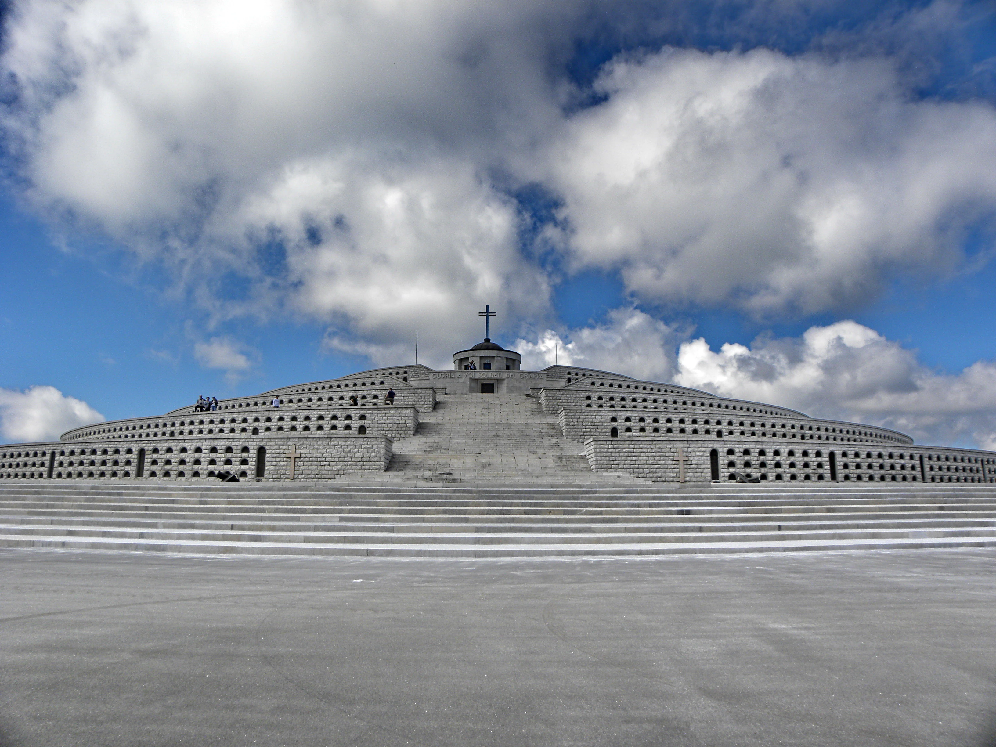 A wide angle, eye level shot of the large, circular, tiered stone Monte Grappa War Memorial set against a bright blue sky with scattered white clouds, featuring a cross at the very top.
