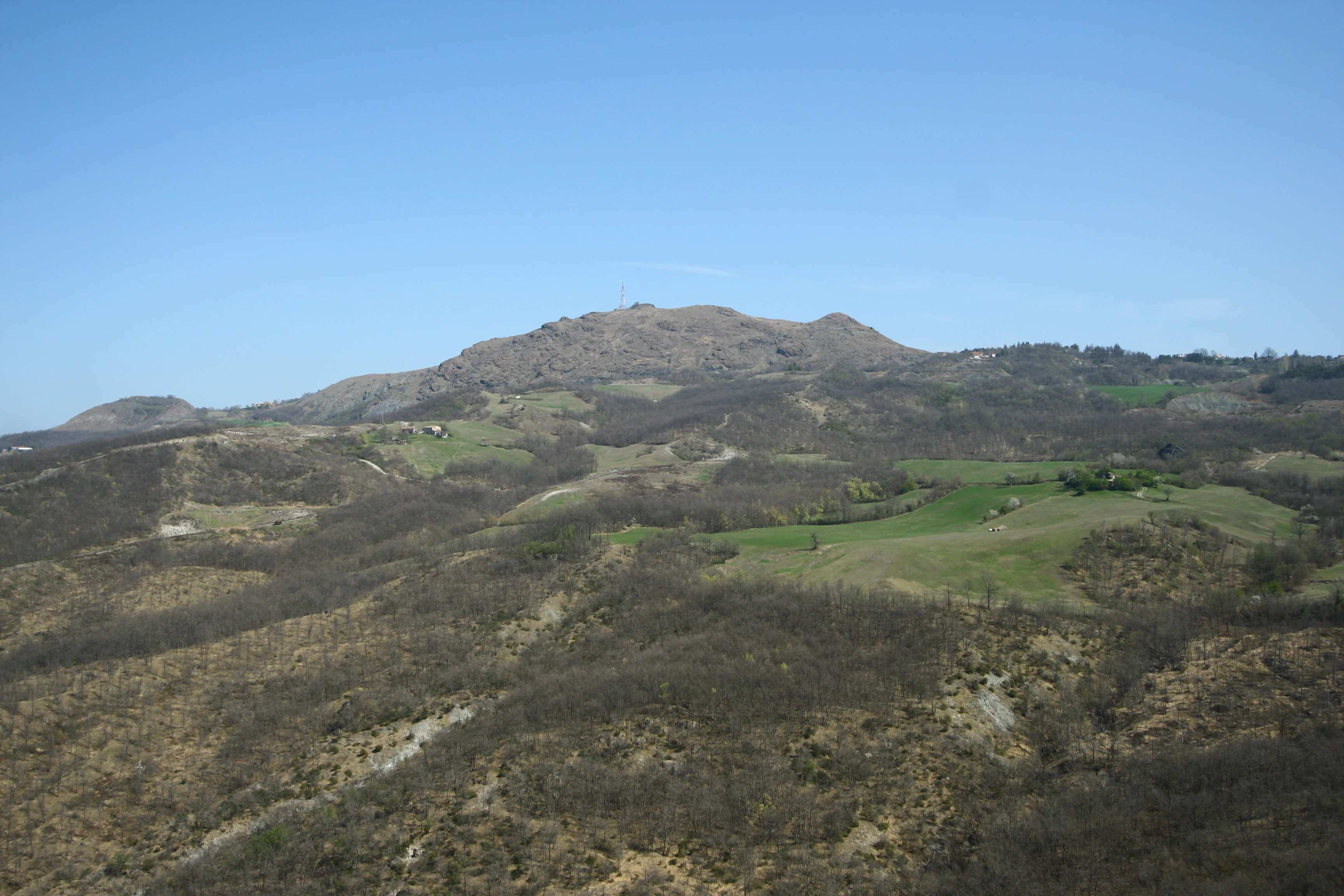 A panoramic mountain landscape view showing the rocky, sparse ridgeline of Monte Prinzera rising above rolling, scrubby hills and patches of green fields under a clear blue sky.