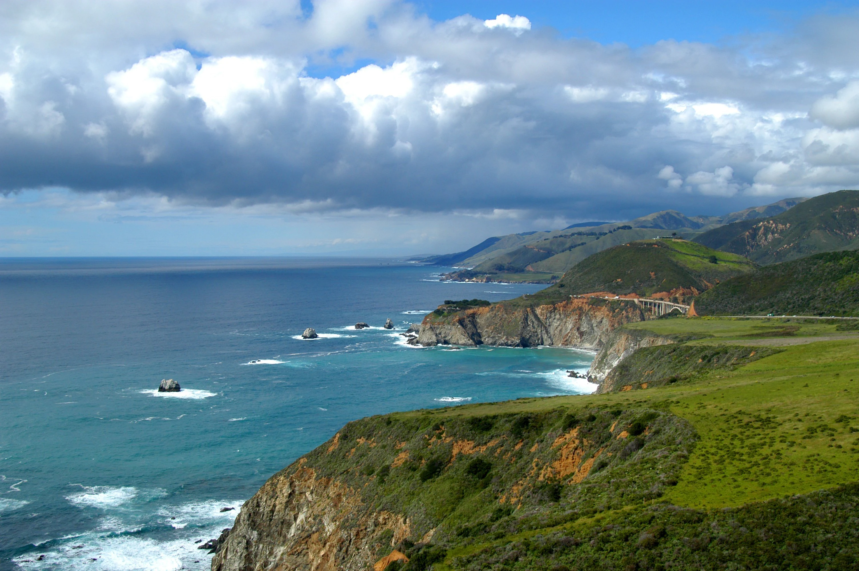 A wide angle view of the California coast, showing green grassy cliffs dropping into the blue Pacific Ocean with the Bixby Canyon Bridge visible in the distance under a partly cloudy sky.