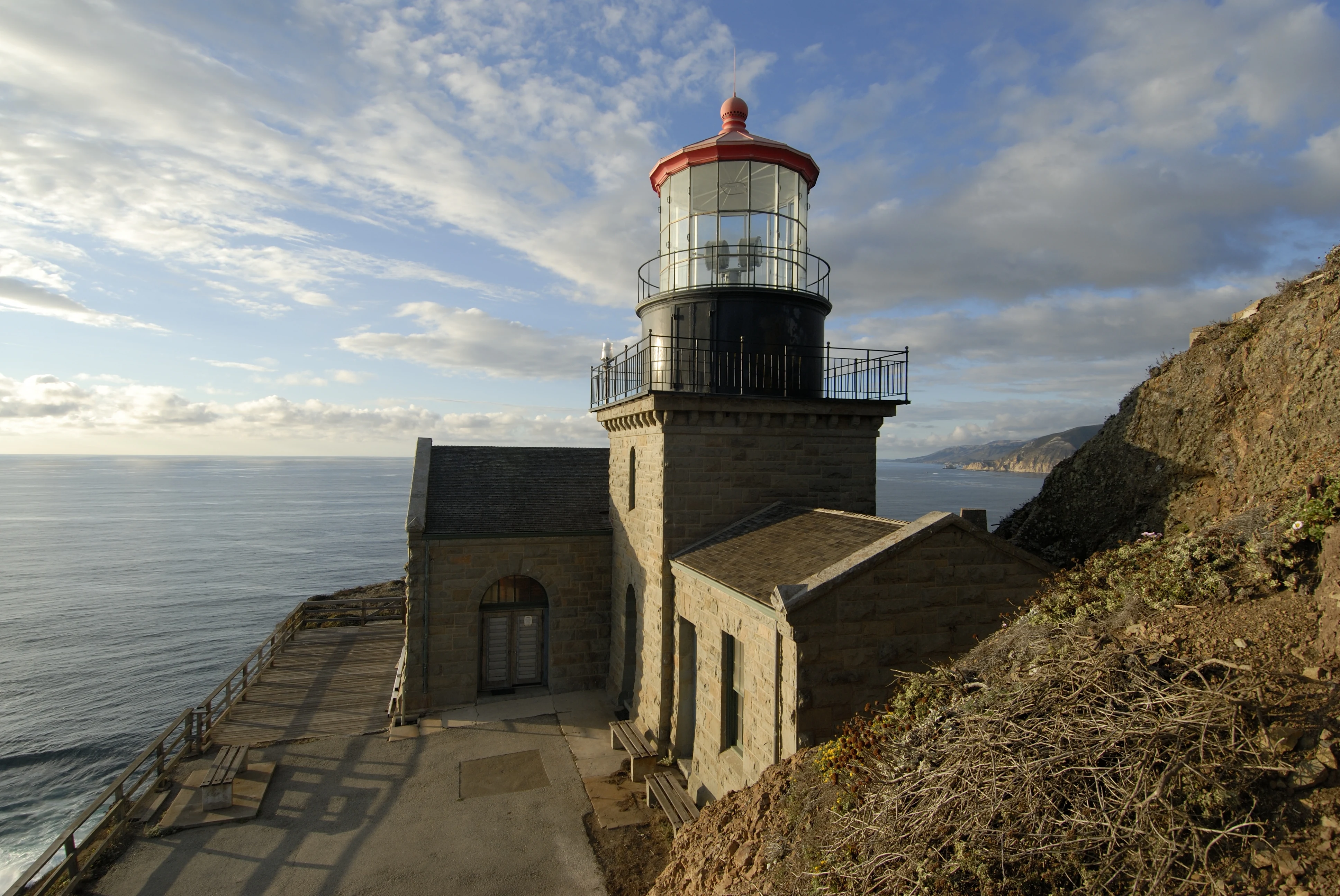 A stone lighthouse with a red-roofed lantern room sits on a cliffside overlooking the Pacific Ocean, with a paved walkway in the foreground and rugged slopes nearby.