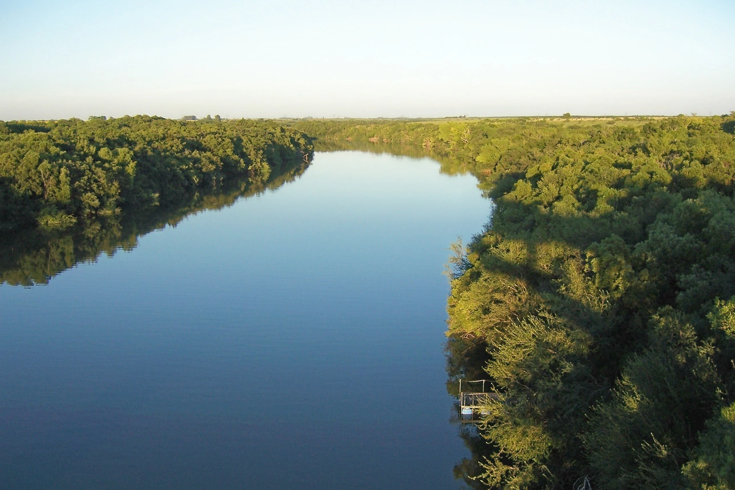 A wide, calm river flows through a dense riparian forest under a soft, golden afternoon light.