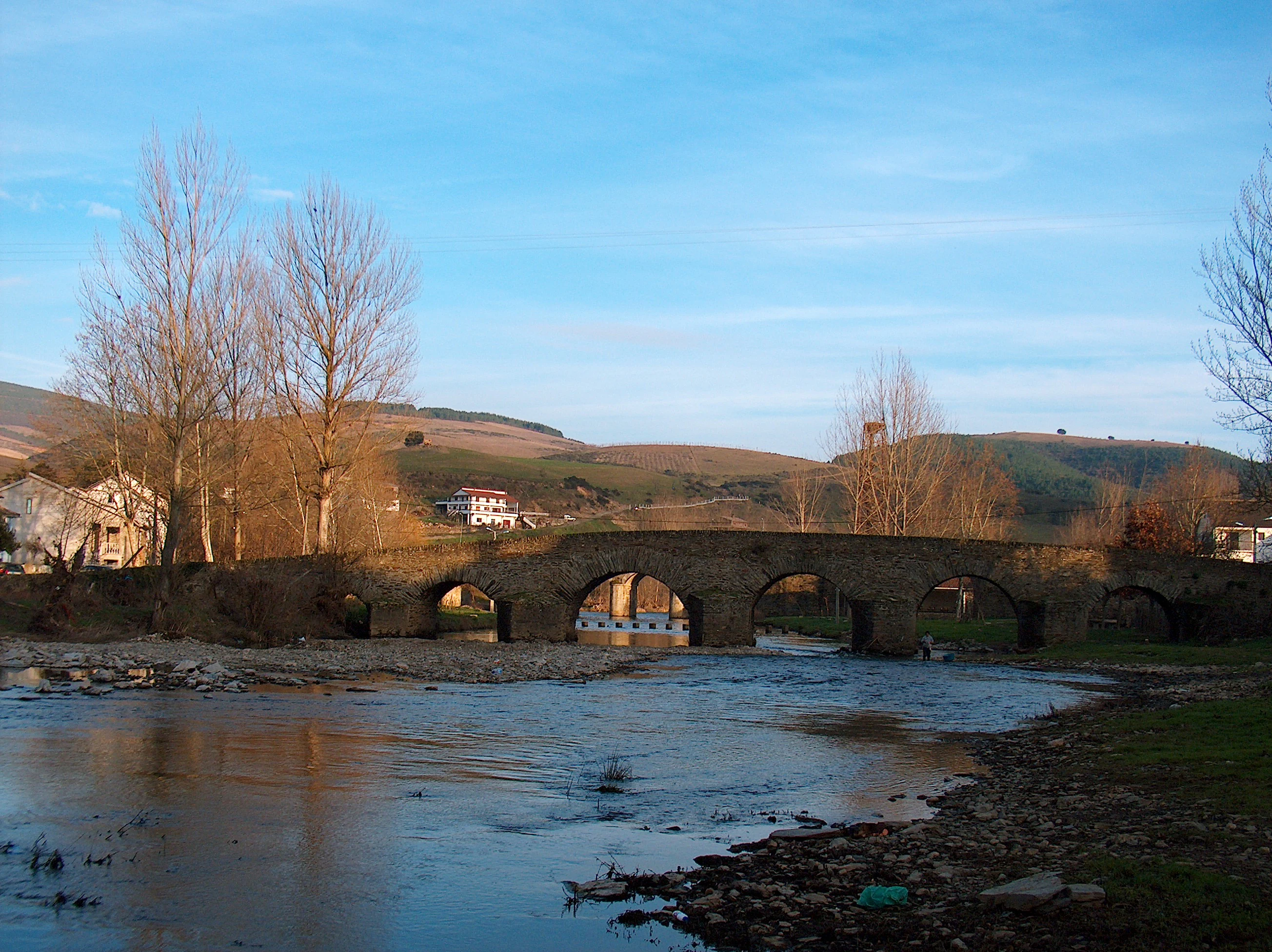 Stone bridge spanning a river with rocky banks and bare trees under a clear blue sky