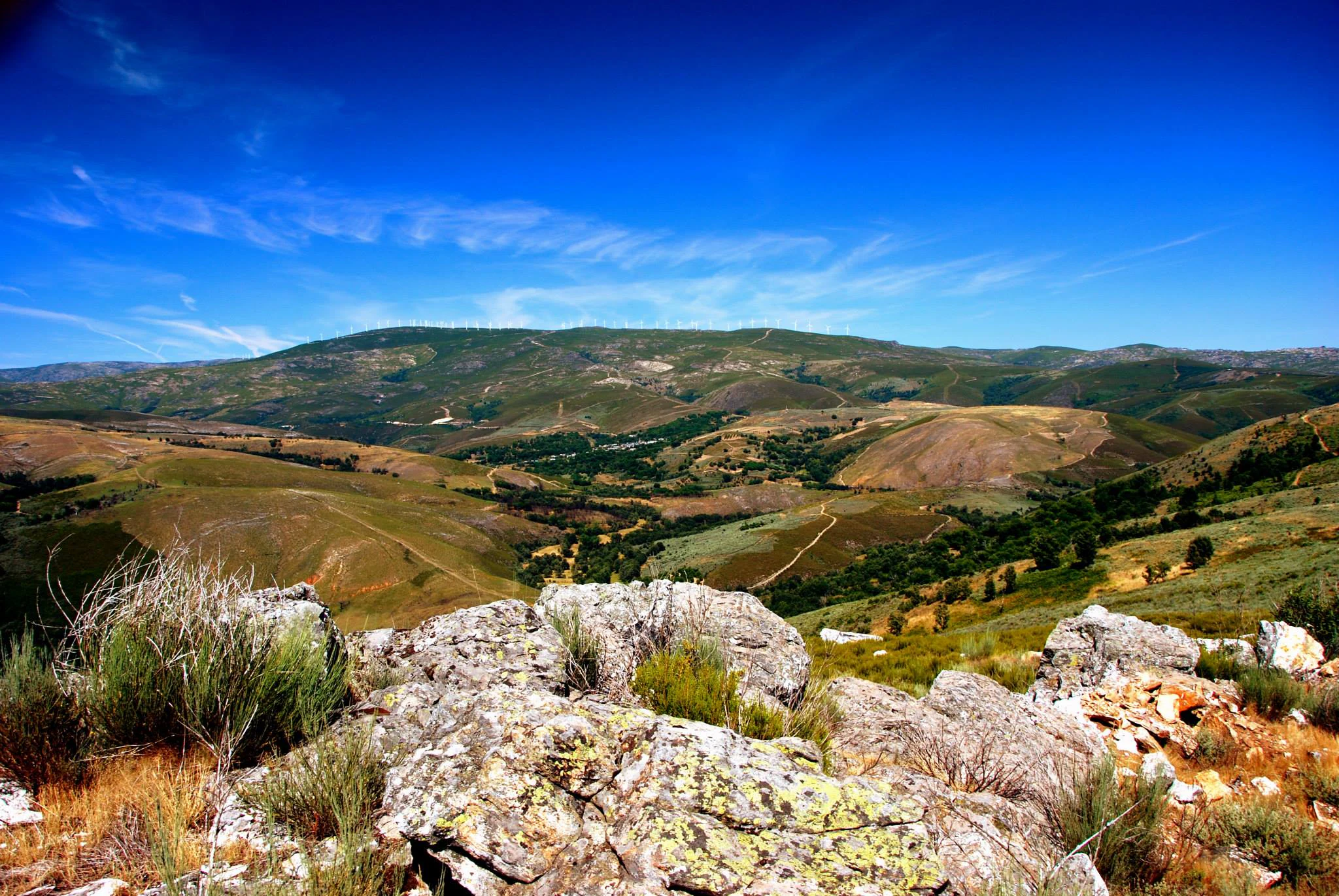Wide landscape view of Montesinho Natural Park showing rocky foreground, grassy hills, and distant mountains under clear blue sky