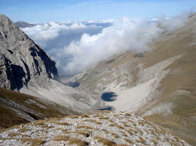 A mountainous landscape with Lake Pilato visible in a valley, surrounded by rocky peaks and clouds under a clear blue sky