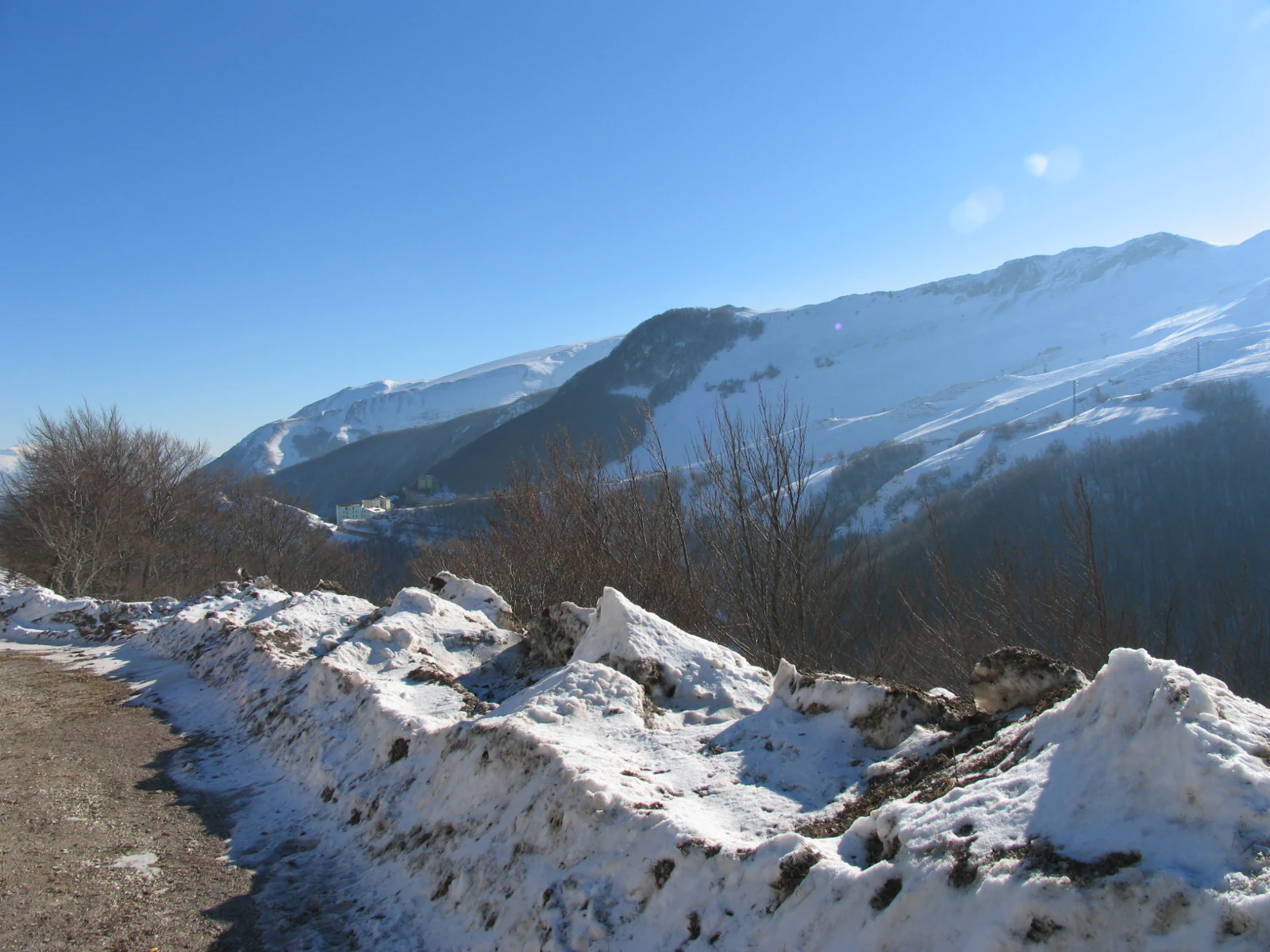 A mountain road with snow piles on the side, snow-covered mountains in the background under a clear blue sky