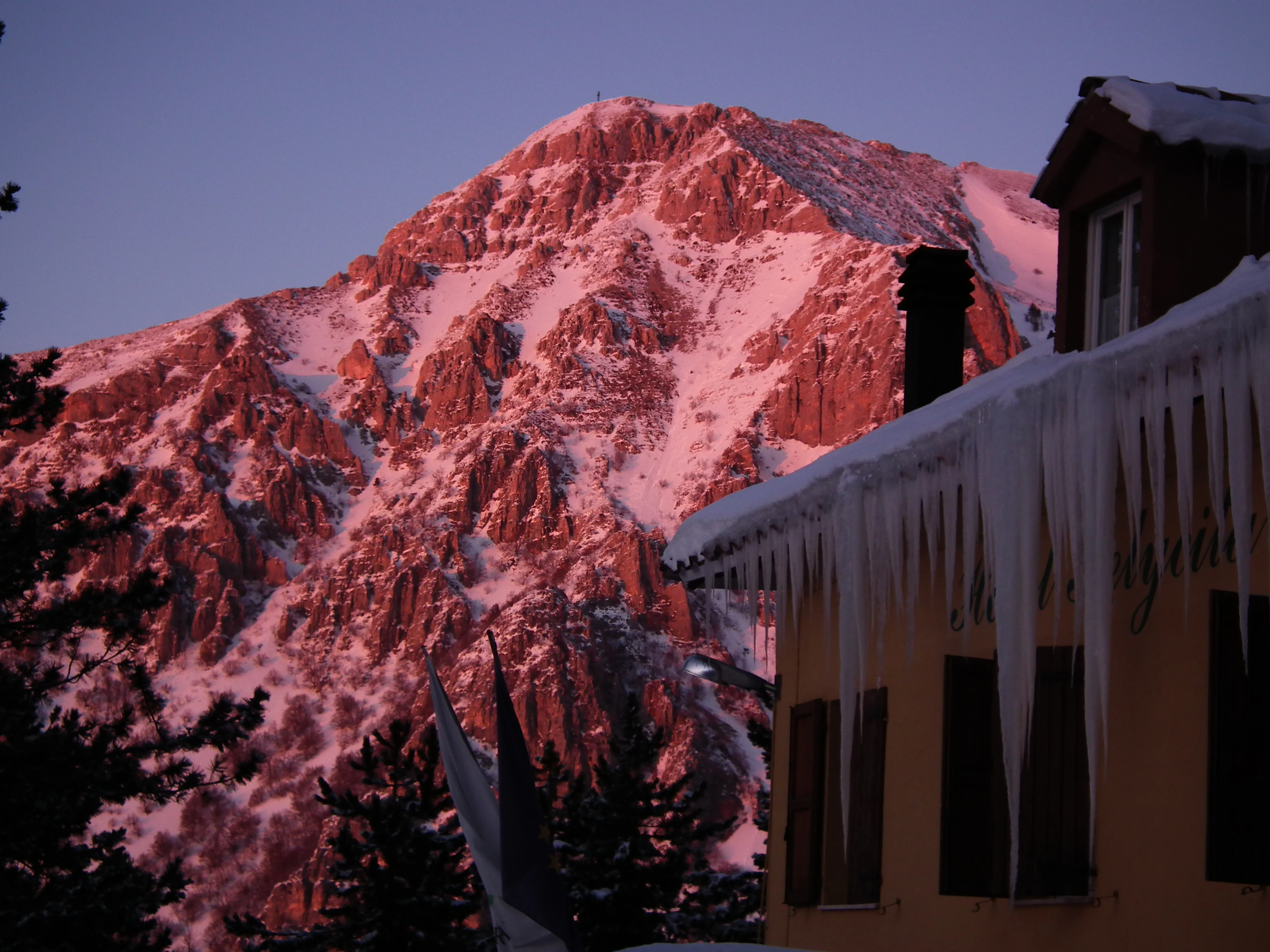 A snow-capped mountain peak with pinkish hues at dusk, a building with icicles on the roof, and trees in the foreground